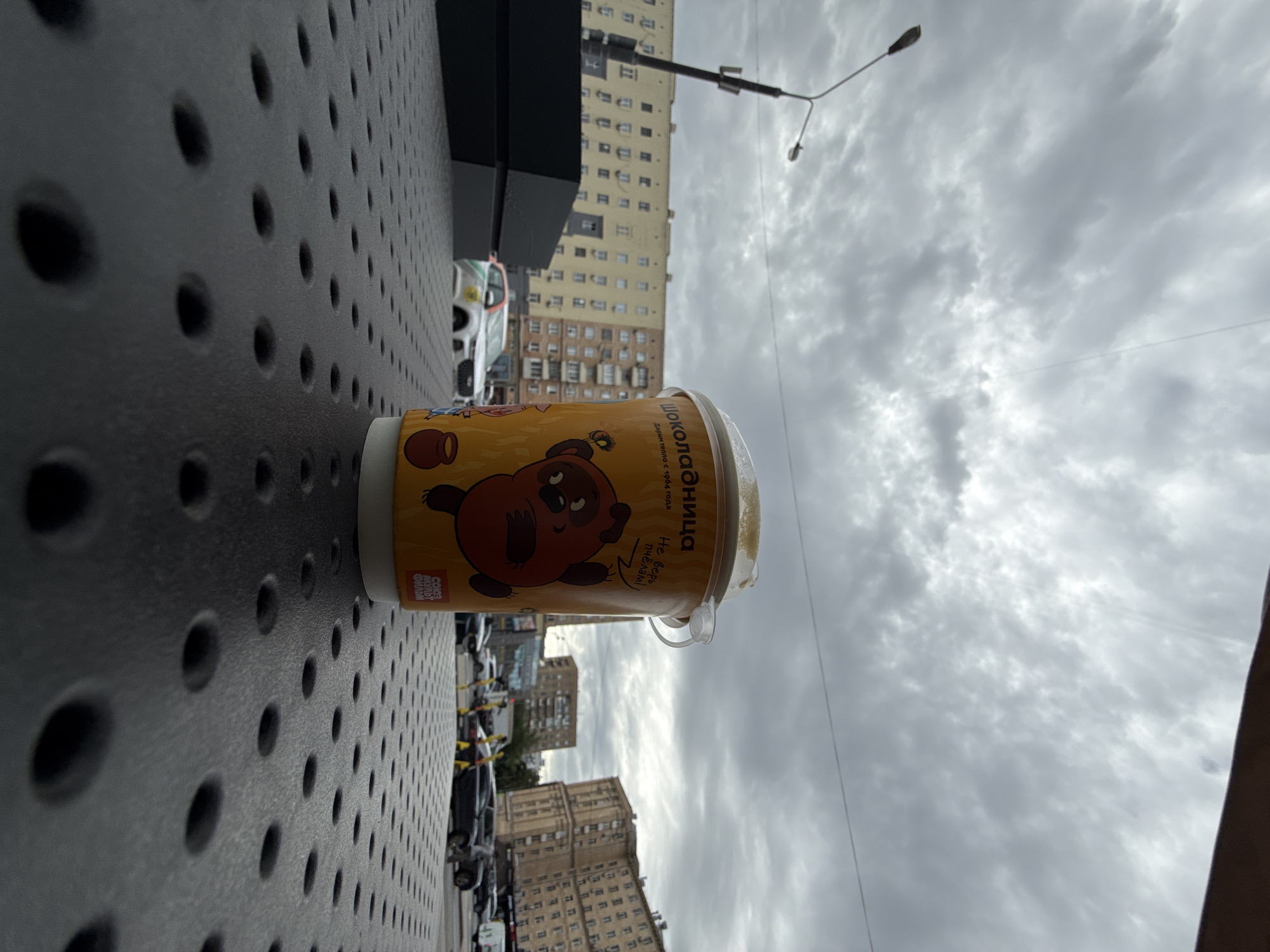 The image presents a cup of "Shokoladnitsa" (chocolate shop) brand hot chocolate with a cartoon bear illustration resting on a perforated metallic surface, likely an outdoor table. The backdrop reveals an urban setting, identifiable as Moscow, Russia, by the architectural style of the tall apartment buildings lining the street and the Russian-language branding on the cup. The sky is overcast, suggesting a cloudy day. Cars are visible parked along the street. A modern street light fixture is seen against the cloudy sky.
