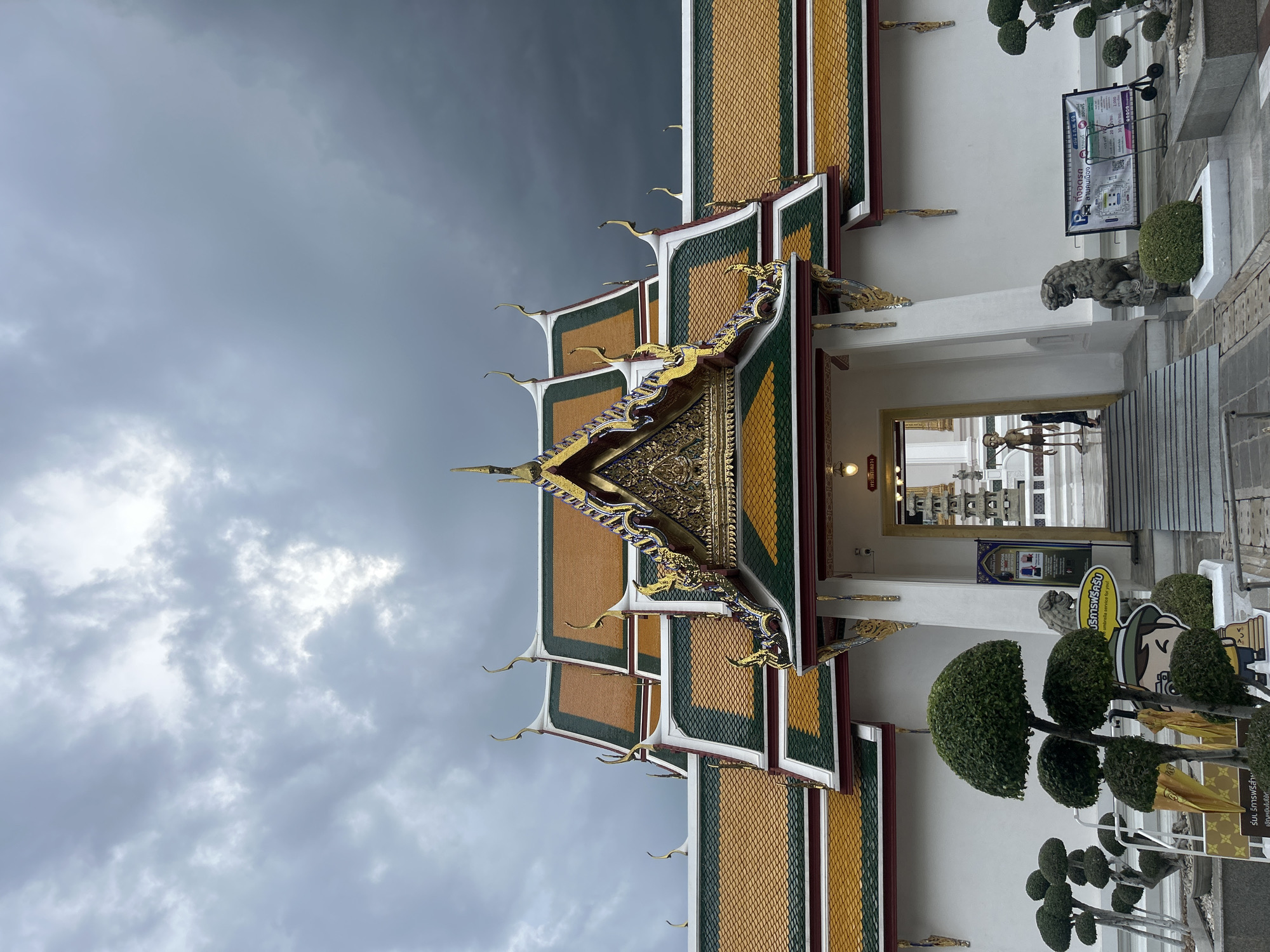 This image shows the entrance to Wat Pho, also known as the Temple of the Reclining Buddha, in Bangkok, Thailand. The temple features traditional Thai architecture with ornate, multi-tiered roofs tiled in orange and green, topped with golden finials. The sky above is overcast and dark, suggesting an approaching storm. In the foreground, there are several artfully trimmed topiaries and a cartoon-like cutout of a person holding a camera and an umbrella. A stone lion statue stands guard near the entrance, and a sign in Thai offers information about parking. Through the entrance, a staircase leads into the temple complex, where a statue of a skeletal figure can be seen. The overall scene conveys a sense of grandeur and cultural significance, with a dramatic sky adding to the atmosphere.
