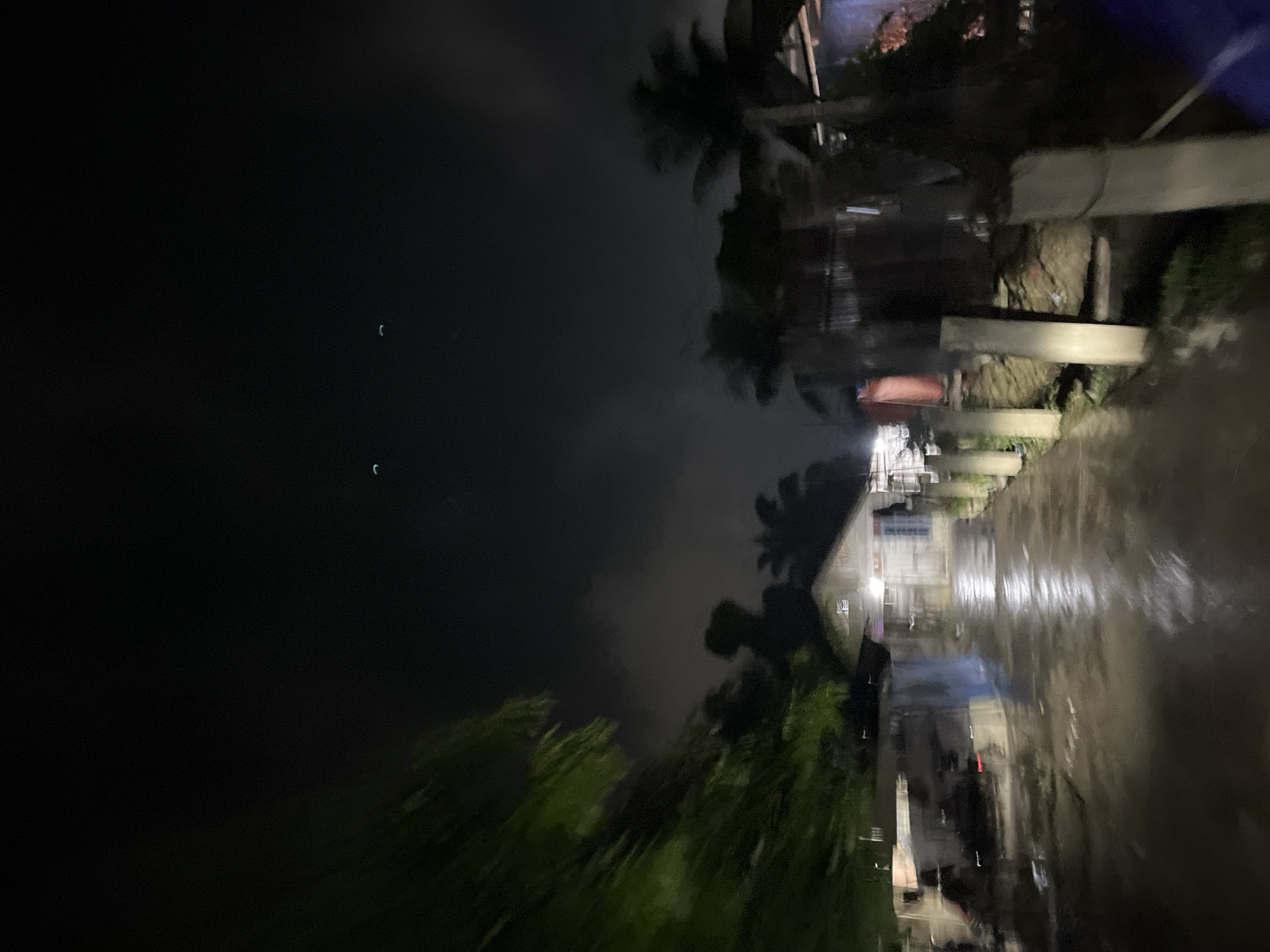 This image depicts a nighttime scene in Benuaraja, Indonesia. The orientation of the photo is difficult to determine, but it appears to be a street or pathway that is wet, suggesting it may have recently rained or is currently raining. The scene is dark, with limited lighting, giving it a mysterious or perhaps serene atmosphere.
In the foreground and extending into the midground, there is a strong sense of motion blur, indicating that the photo was likely taken from a moving vehicle or with a fast-moving subject. This blur obscures many details, but glimpses of greenery, possibly trees or foliage, can be seen.
On the right side of the image, the reflection of lights on the wet surface creates a distorted, elongated effect. Buildings or structures are visible along this side, though their details are also softened by the darkness and motion. Some of these structures appear to be simple dwellings.
In the far left of the image, against the dark sky, there are two faint, crescent-shaped lights, which could be the moon or possibly distant lights. The sky itself is dark and appears cloudy, adding to the nighttime ambiance.
The overall impression is one of a quiet, perhaps humid night in a tropical location, possibly a village or rural area in Indonesia, characterized by wet surfaces and a feeling of movement. There is no visible text in the image.