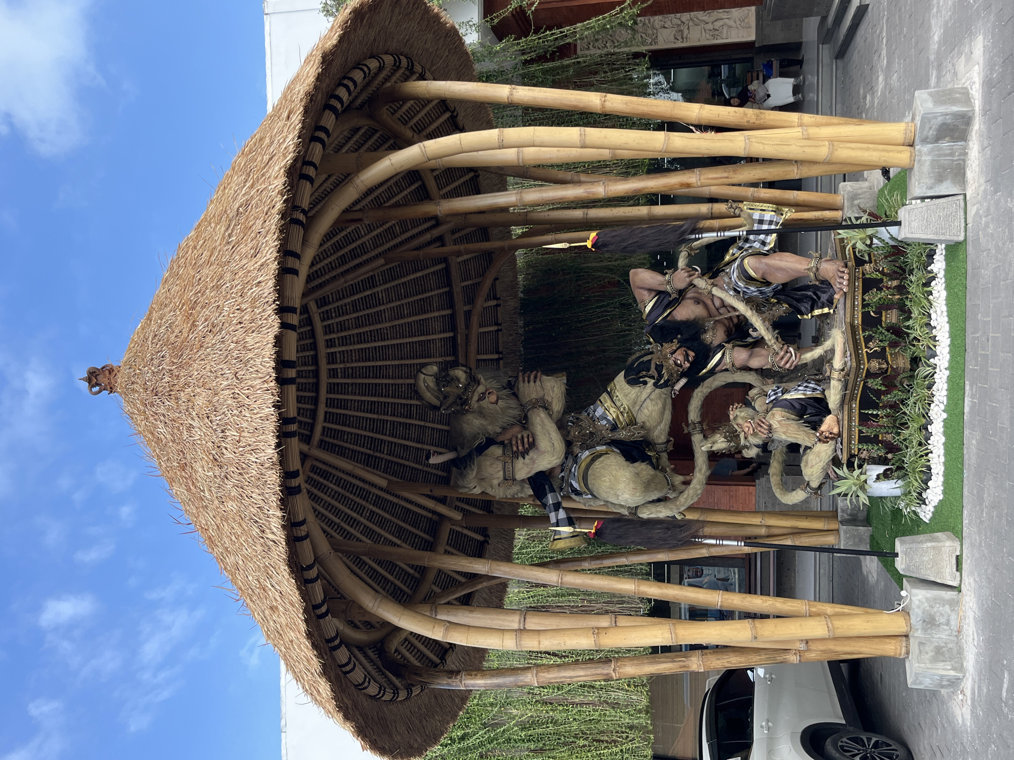 The image shows a thatched-roof pavilion with bamboo supports, housing a sculpture of several figures. The figures appear to be human-like with light-colored, hairy skin and are dressed in traditional Balinese attire, including black and gold patterned clothing and checkered loincloths. One figure is depicted in a dramatic pose, holding a staff. The sculptures are intricately detailed, with visible jewelry and facial features. The setting is outdoors, with a paved walkway and lush greenery surrounding the pavilion. In the background, a building with intricate stone carvings is visible. A white car is parked in front of the pavilion, and a bright blue sky with some white clouds suggests it is daytime. The scene is likely a cultural display or a religious monument, possibly in Kuta, Indonesia, given the context. The overall impression is one of cultural richness and artistic craftsmanship.