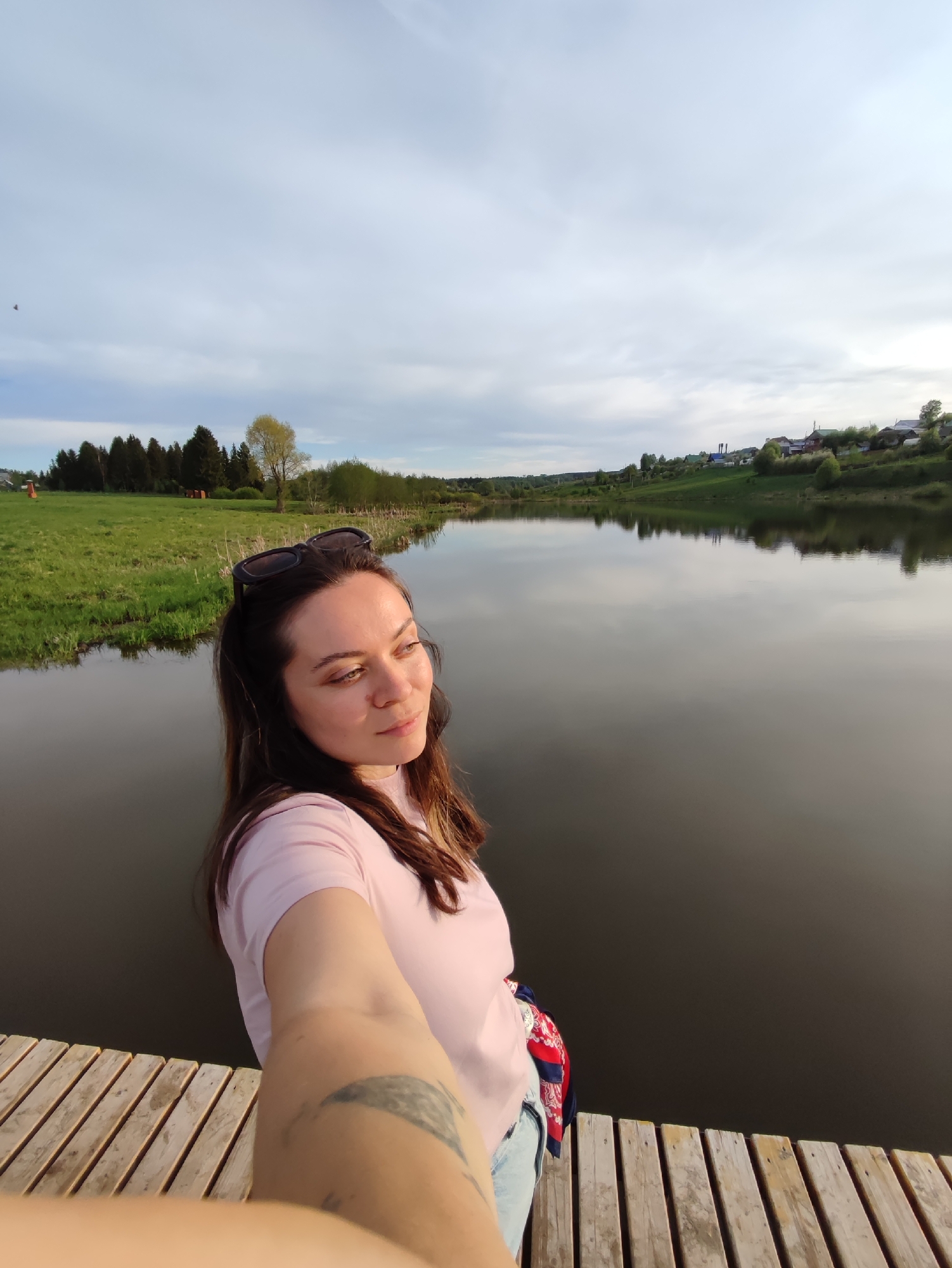 A young woman takes a selfie on a wooden dock in Фоки, Russia. She wears a light pink T-shirt, sunglasses on her head, and a colorful scarf around her waist. Her left arm, visible in the foreground, has a tattoo. A calm, dark body of water extends behind her, reflecting the sky and surrounding landscape. On the left bank, there is green grass and a stand of trees. The right bank is a gentle slope with scattered houses and vegetation. The sky is overcast with a mix of gray and white clouds.