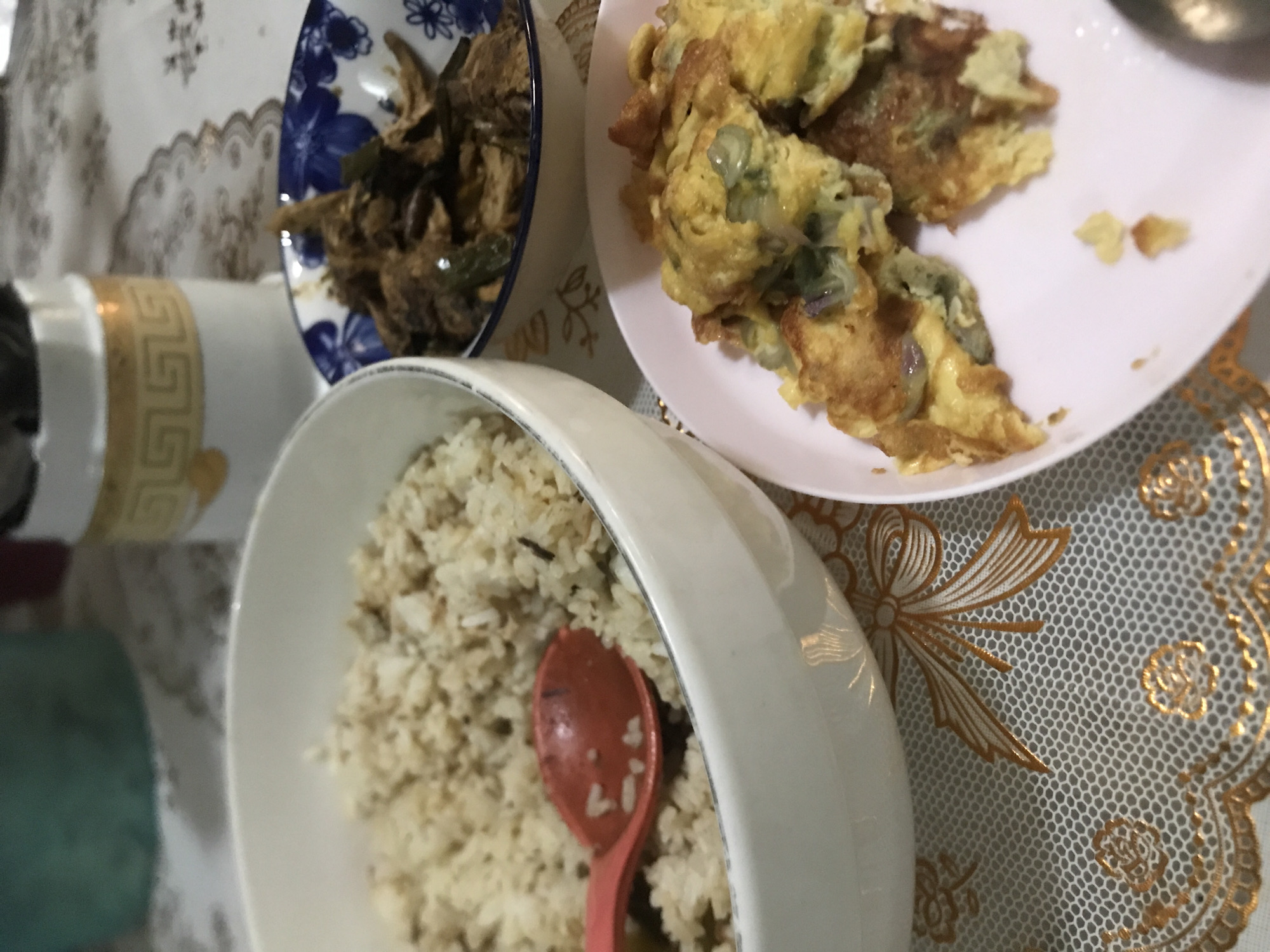 The image shows a meal on a table. There's a bowl of rice with a pink spoon in it, a blue and white patterned bowl containing a dark-colored dish, and a pink plate holding what appears to be scrambled eggs or an omelet. The meal is set on a tablecloth featuring a pattern. The exact location within Lhokseumawe, Indonesia is not discernible from the provided image.