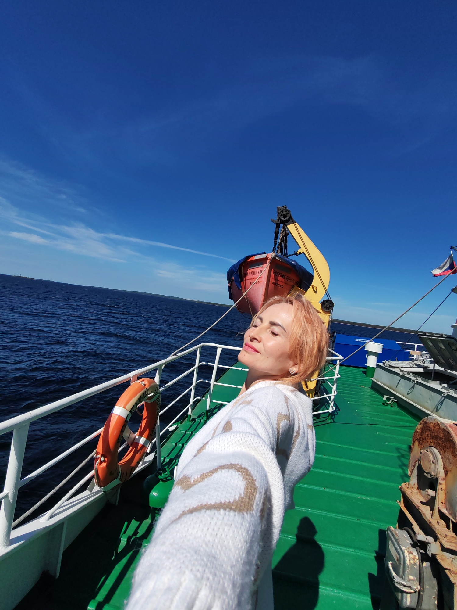A woman with short, light-colored hair is taking a selfie on the deck of a ship. She is wearing a white sweater with a patterned design. Her eyes are closed, and she appears to be enjoying the sunlight. To her left, a life preserver is attached to the railing. In the background, there is a lifeboat suspended by a crane, and a Russian flag flies from a mast. The ship is on a body of water, likely the White Sea, with a landmass visible in the distance. The deck of the ship is painted green, and there are various pieces of equipment, including a rusty winch. The sky is a clear, bright blue with a few wispy clouds. The overall scene suggests a bright, sunny day at sea. The location is identified as Shal'skii, Russia.