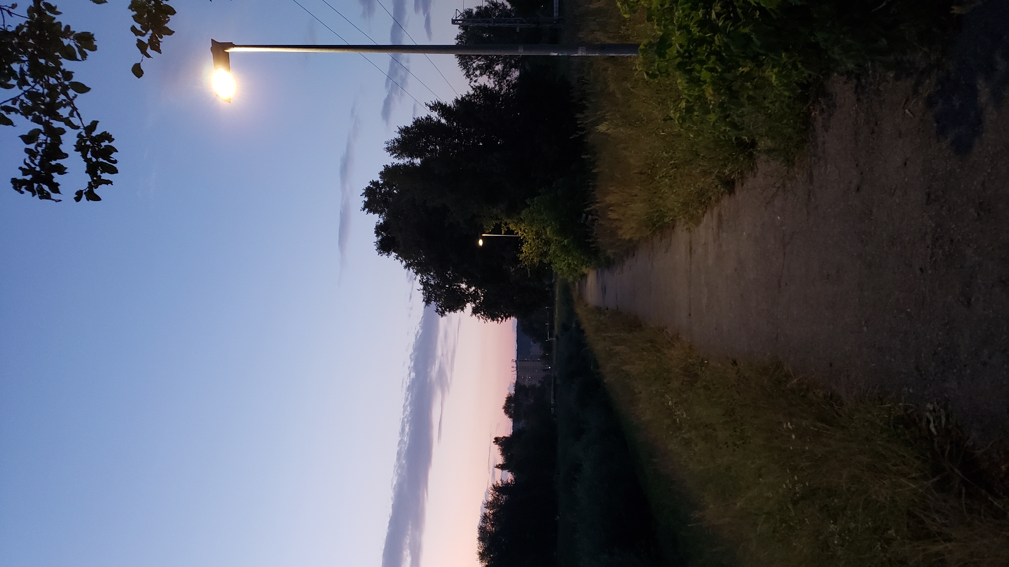 A pathway through a grassy area leads towards a distant city skyline at dusk. A solitary streetlight illuminates the path and the surrounding vegetation. The sky displays a gradient of blue and pink hues, with a few wispy clouds. In the background, silhouetted trees and buildings are visible. Power lines are strung between poles, and a distant building with multiple windows stands out against the twilight sky. The scene appears to be located in the city of Prešov, Slovakia, possibly near a park or a riverbank.