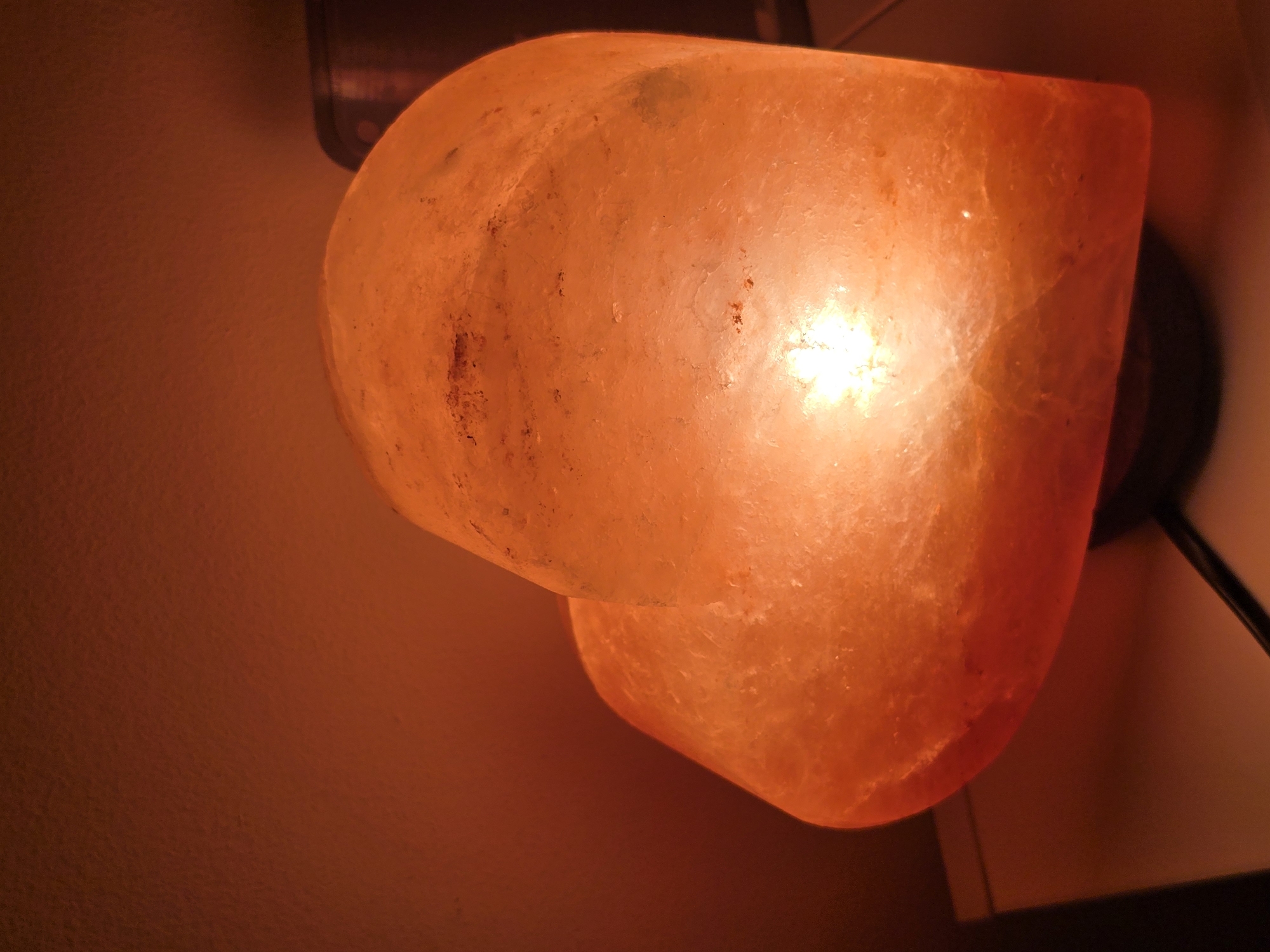 A heart-shaped Himalayan salt lamp sits on a table, casting a warm, orange glow. The lamp's surface is rough and crystalline, with natural imperfections and variations in color. The background is a blurred, warm-toned wall, suggesting an indoor setting. The lamp is illuminated from within, highlighting its translucent quality. There are no people or animals visible in the image, nor any indications of the specific location within the city of Golyanovtsi, Bulgaria. The scene is static and peaceful.