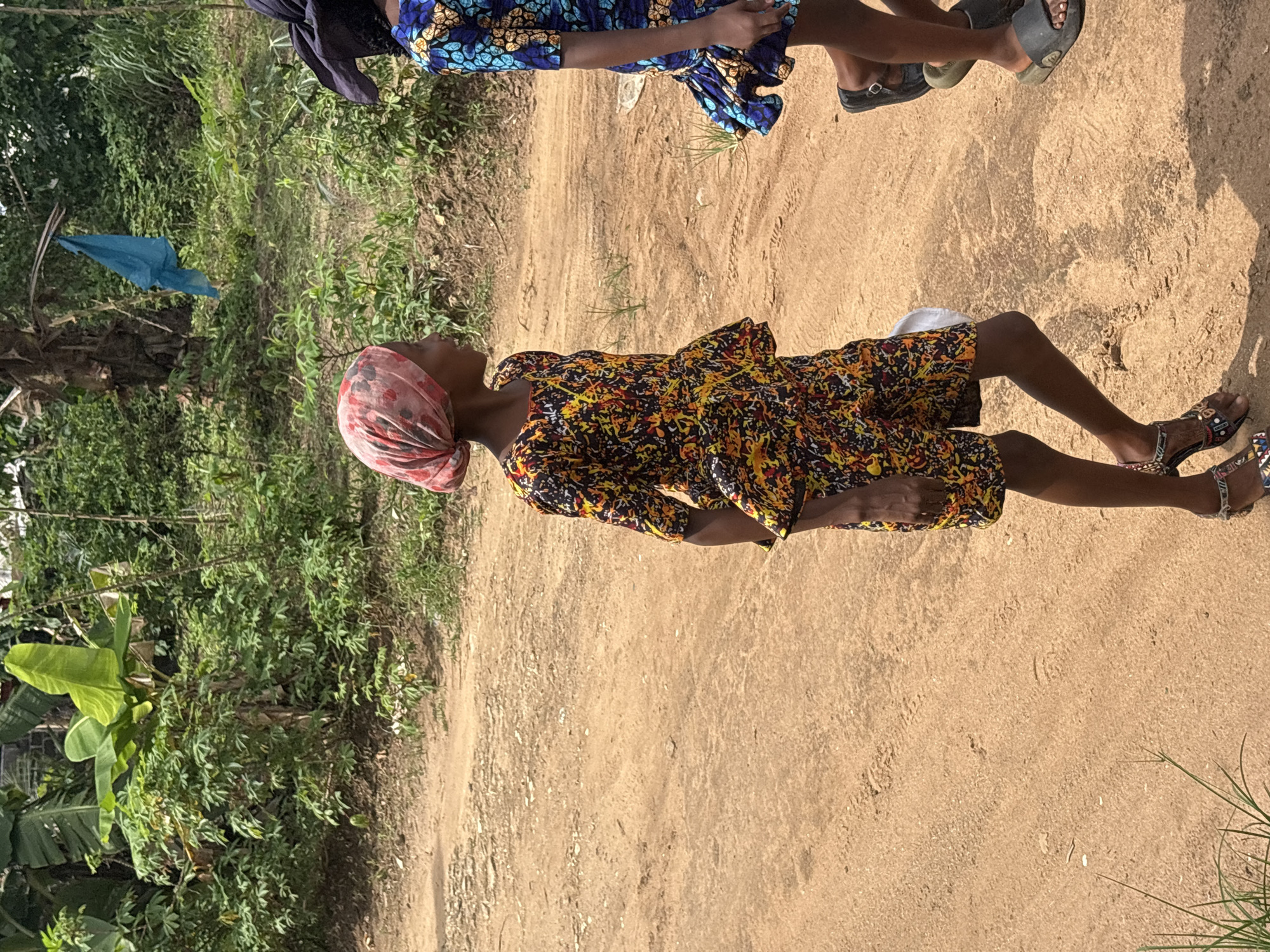The image shows a dirt road in what appears to be a rural area near Ikot Ekpene, Nigeria. A woman wearing a floral print dress and a pink headscarf is standing on the road. Another person wearing a blue floral print dress and a dark head covering can be partially seen at the top of the image. The ground is dirt and there is vegetation, including banana trees and other green plants, on the left side of the frame. A blue object, possibly a piece of cloth, is hanging from a tree branch in the background.