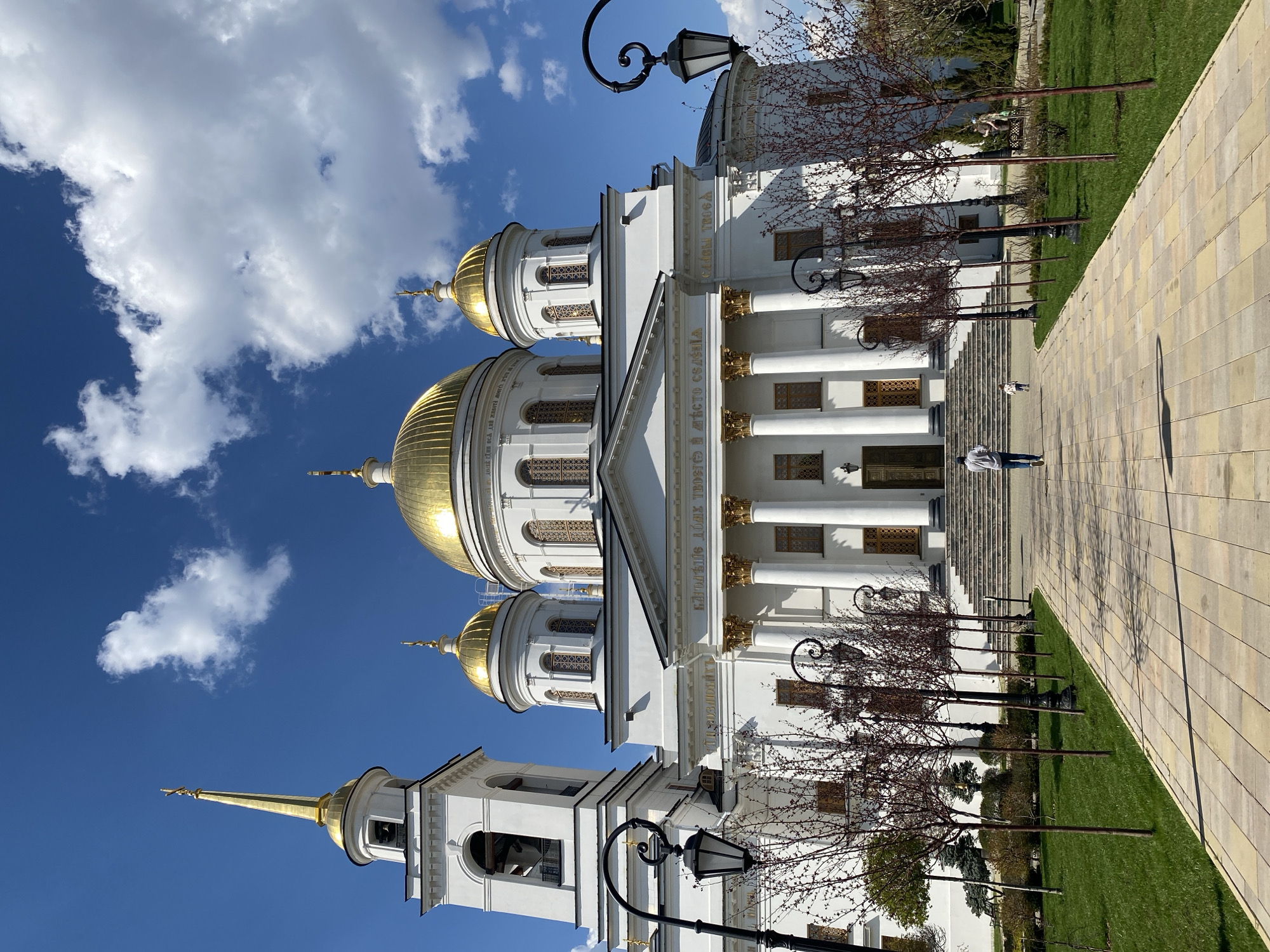 The image shows the Church on Blood in Yekaterinburg, Russia, viewed from a ground-level perspective. The white church features prominent gold domes and classical architectural elements. A paved walkway runs along the right side of the frame, bordering a strip of grass. Several bare trees line the walkway, interspersed with black lampposts. Two people are on the stairs in front of the church, with one person walking towards the building. A blue sky with scattered white clouds is visible above the church.