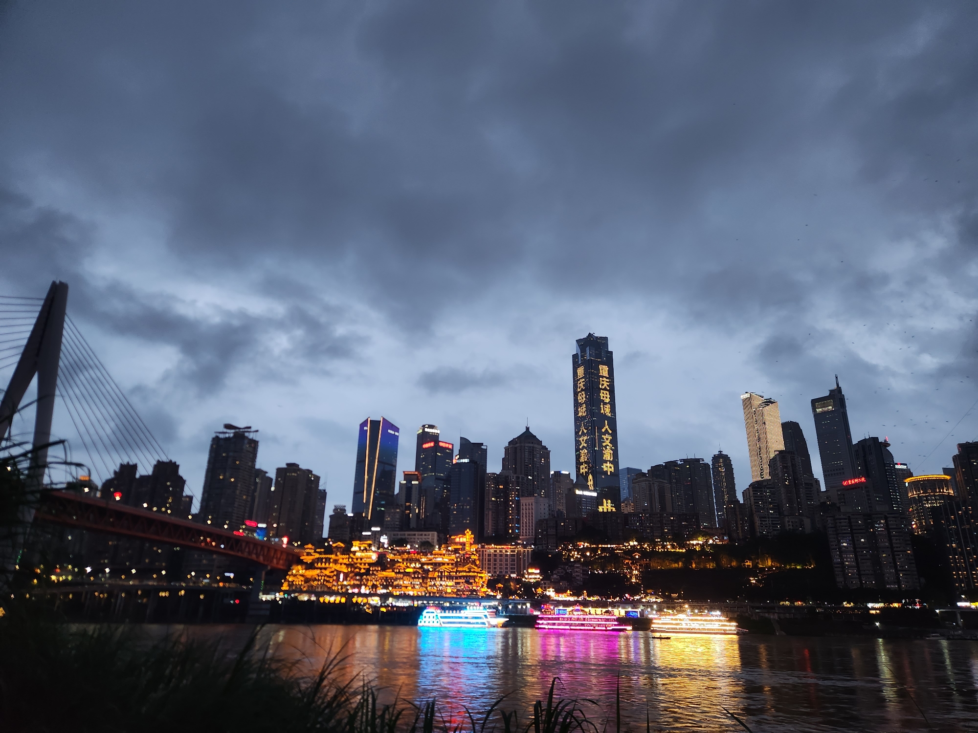 The image captures a cityscape at dusk in 江北城街道, China. In the foreground, water reflects the lights of the city. A bridge with a large support tower is visible on the left. In the middle ground, a cluster of illuminated buildings creates a skyline, interspersed with modern skyscrapers, some bearing Chinese characters. Several brightly lit boats float on the water. The sky is filled with dark, overcast clouds.
