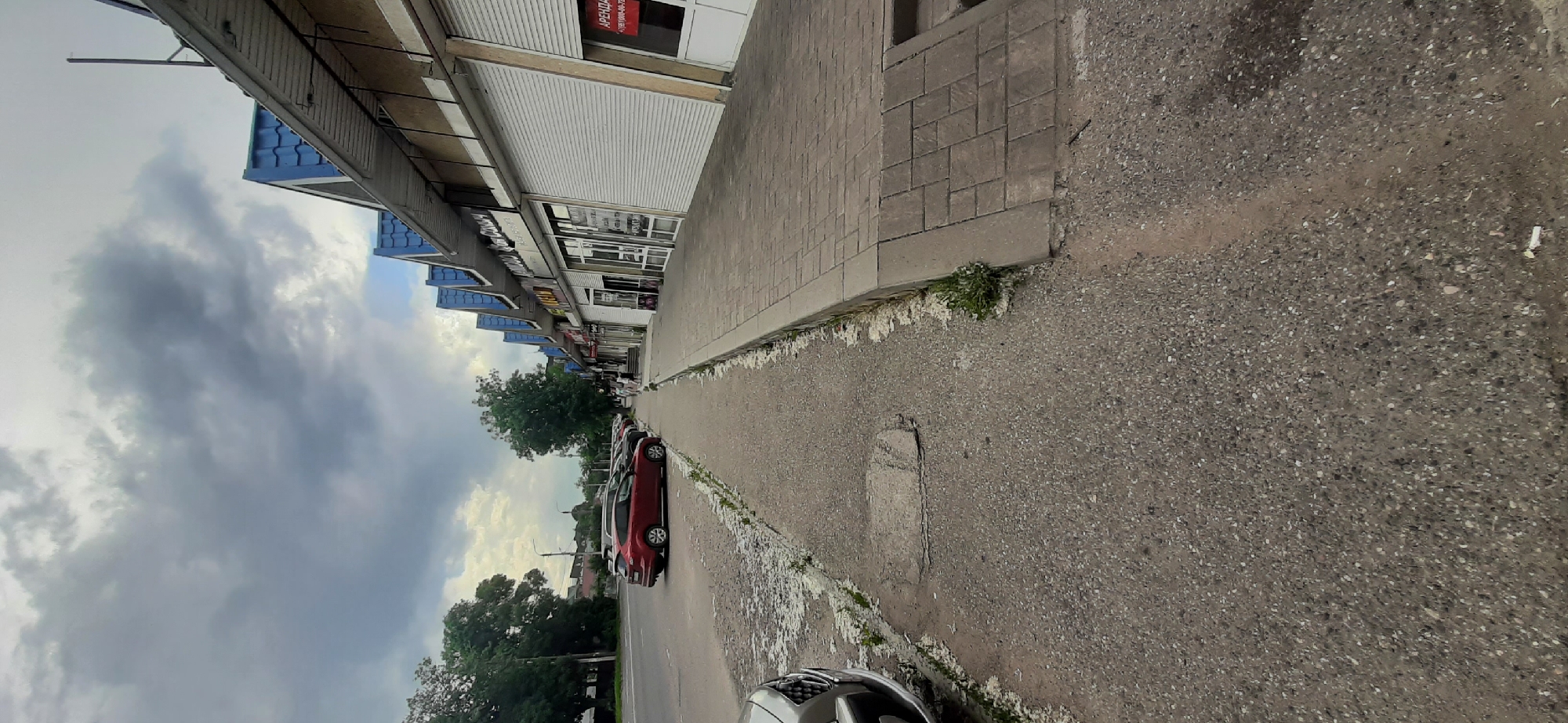 This is a landscape photograph of a street scene in Yessentuki, Russia. The image is oriented vertically, with the sky taking up the left half of the frame and the street and buildings on the right. The sky is filled with dramatic, dark clouds, suggesting overcast or stormy weather.
The right side of the image features a row of buildings with storefronts. Many of the storefronts have rolling metal shutters, with some of them partially or fully closed. One storefront has a red sign with white text that appears to be an advertisement or a "for rent" notice. The buildings have blue awnings over them. In front of the buildings is a sidewalk made of gray paving stones.
Along the edge of the sidewalk, bordering the street, is a strip of rough, unkempt ground with some sparse vegetation and white debris. The street itself is paved with asphalt and shows signs of wear, with some patches and cracks. Several cars are parked along the side of the street, including a red car prominently visible in the foreground.
The overall impression is of a somewhat utilitarian, possibly older, commercial area. The cloudy sky and the presence of parked cars and closed shutters do not suggest a particularly busy or cheerful atmosphere, but rather a common, everyday scene. There are no people clearly visible in the image. The time of day is difficult to ascertain precisely due to the overcast sky, but it appears to be daytime.