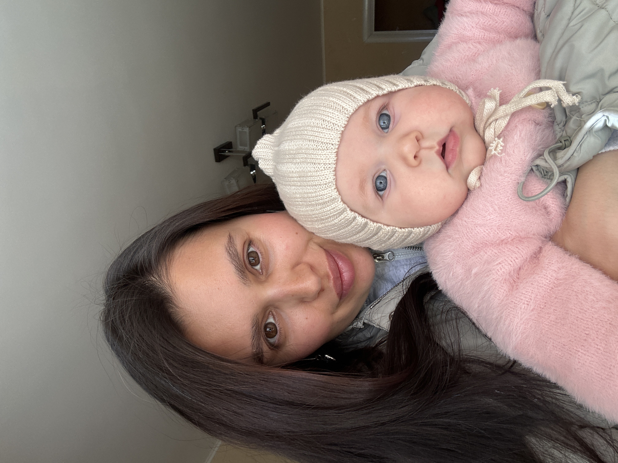 A woman with long, dark hair is holding a baby. The baby has blue eyes and is wearing a white knitted hat and a pink fuzzy jacket. The woman is wearing a gray jacket. The background is a plain wall with a light fixture. The location is Chaikovsky, Russia.