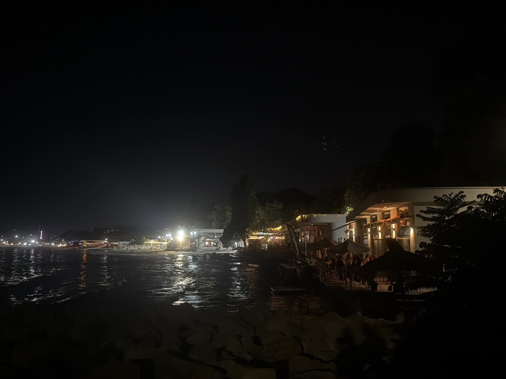 The image depicts a night scene along the coast of Varna, Bulgaria, likely at the central beach area. The foreground is dominated by large, dark rocks, suggesting a breakwater or natural shoreline. Beyond the rocks, the sea is visible, reflecting the lights from the shore and illuminated by moonlight or ambient light, creating a shimmering effect on the water's surface.
Along the coastline, illuminated by numerous lights, are several establishments, likely restaurants or beach bars. These buildings feature bright lights and strings of decorative lights, casting warm glows onto their surroundings. Patrons can be seen seated at tables, suggesting a lively atmosphere. Umbrellas are deployed in front of some of these establishments, indicating outdoor seating areas.
Further down the coast, more lights are visible, illuminating buildings and possibly a Ferris wheel, suggesting a more distant part of the boardwalk or amusement area. The sky is dark, with a few faint clusters of lights, possibly stars or distant city lights. The overall impression is of a vibrant, illuminated waterfront scene at night.