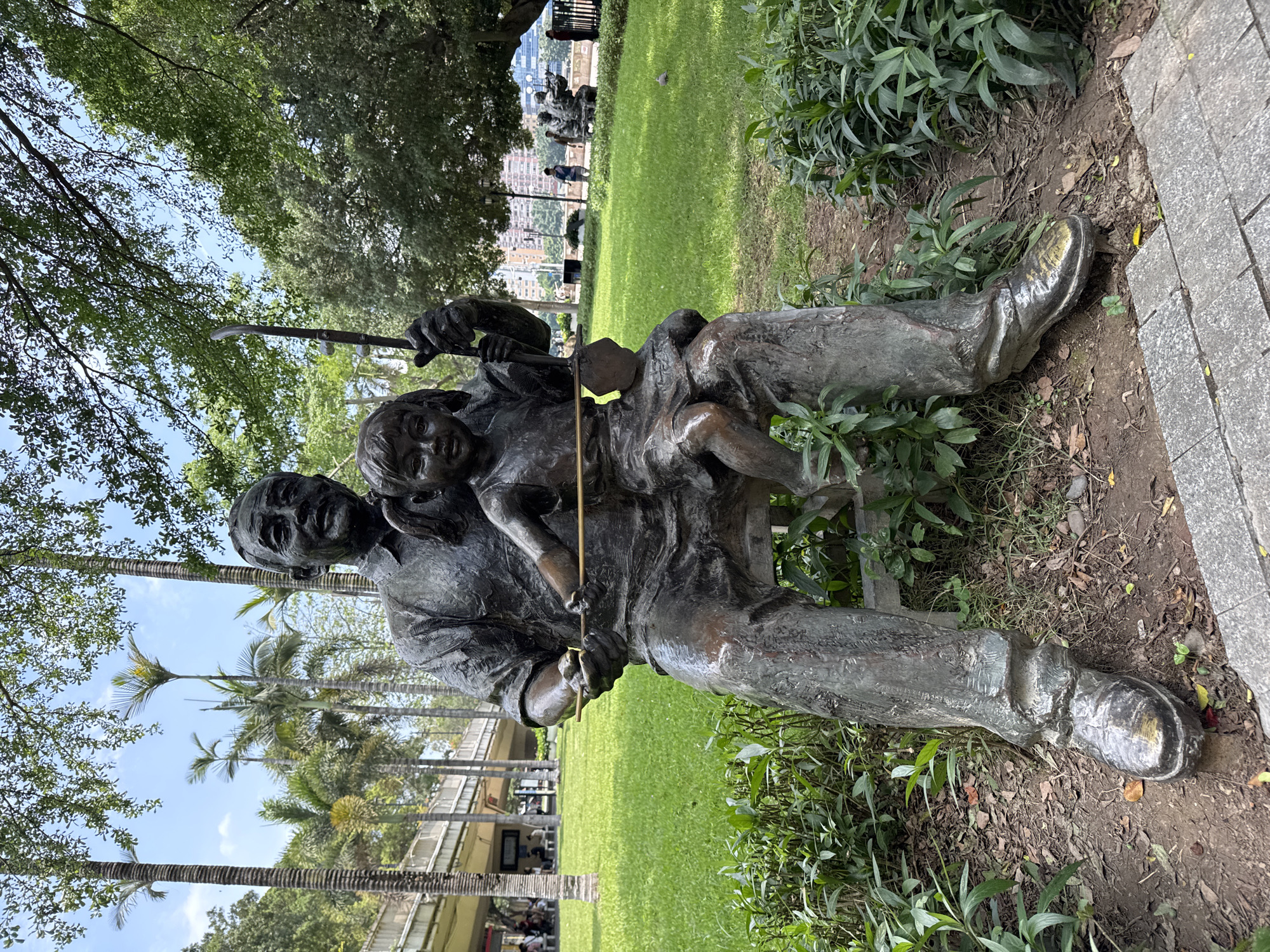 A bronze statue depicting an older man seated with a young child on his lap sits in a park area in Liwan District, China. The statue is positioned next to a paved walkway with a patch of dirt covered in vegetation and dead leaves. The man is holding an Erhu and the child is smiling. In the background is a green grassy area, trees, and other park visitors. There is a building visible in the distance. Palm trees are also seen in the area.