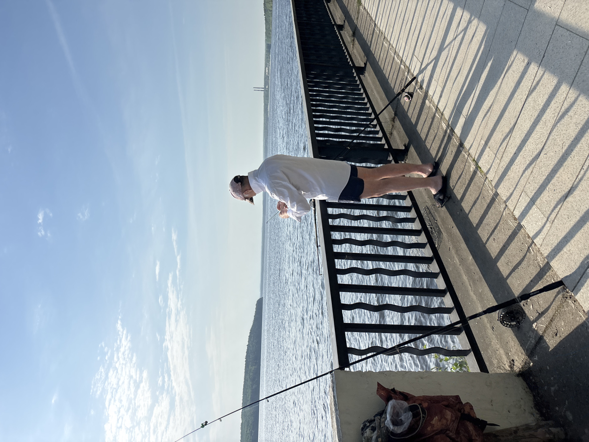 A man is fishing off a concrete pier, possibly in Chaikovsky, Russia. He is wearing a white shirt, blue shorts, and a cap, leaning over a black metal railing to look at the water. Two fishing poles are positioned near him, one closer to his feet and another further down the pier. A brown bag is near the railing. The water stretches out into the distance, with a wooded shoreline visible on the left. The sky is bright blue with some scattered clouds. The pier's surface has a pattern of shadows.