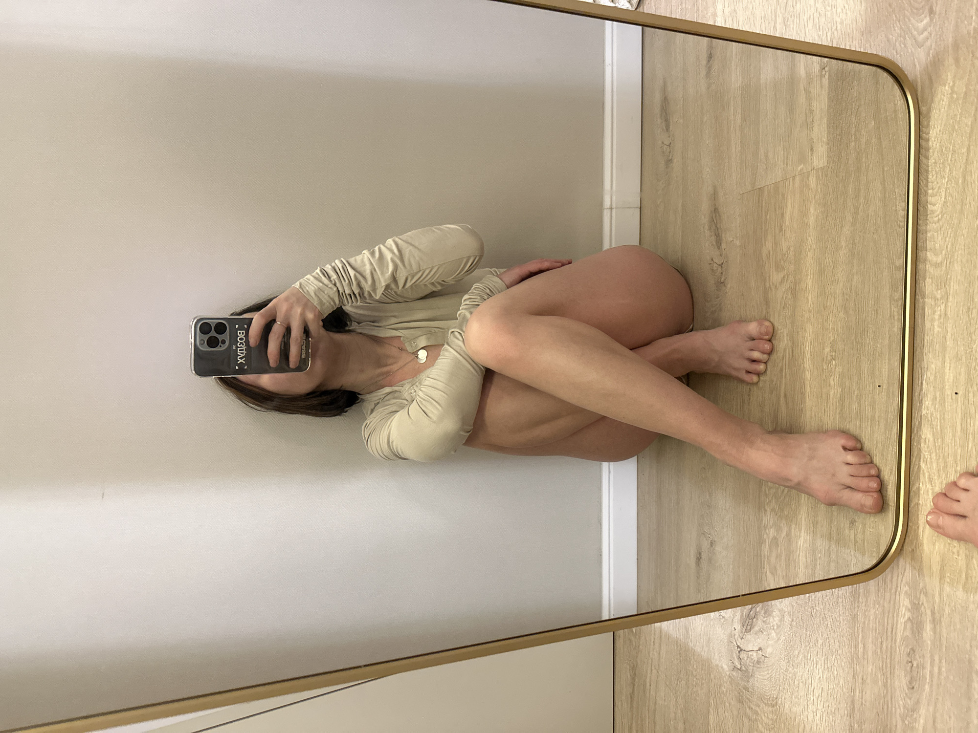 A person is seen taking a selfie in a mirror. The person is lying on a light-colored wooden floor against a light-colored wall, wearing a light-beige long-sleeved top. The phone case is black and has some writing on it. There is a gold-framed mirror showing the person and the environment. The location is not specified as being in Votkinsk, Russia.