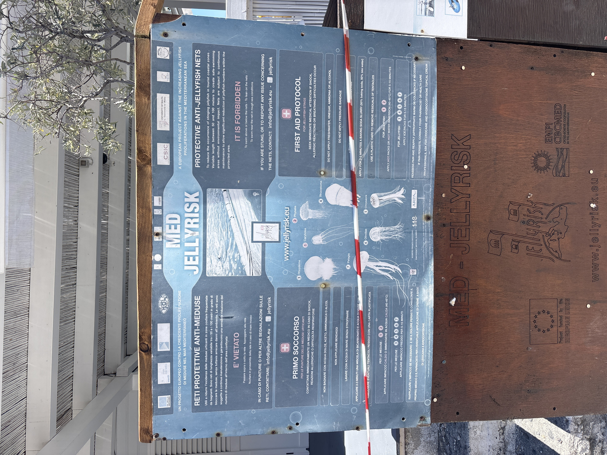 The image shows a sign in Lipari, Italy, providing information about jellyfish and protective anti-jellyfish nets in the Mediterranean Sea. The sign, predominantly in Italian and English, warns against swimming above or below the nets and leaning on them. It includes a first-aid protocol for jellyfish stings, advising not to apply freshwater, vinegar, ammonia, or alcohol and recommending using a plastic card to remove tentacles and applying hot or cold packs. The sign also displays logos of the European Union, ENPI CBCMED, CSIC, and the University of Malta, indicating that the project is funded by the European Union. The sign is attached to a wooden frame near some white slats. The jellyfish illustrations include Carybdea, Physalia, Rhopilema, Pelagia, Olindias, and Chrysaora.