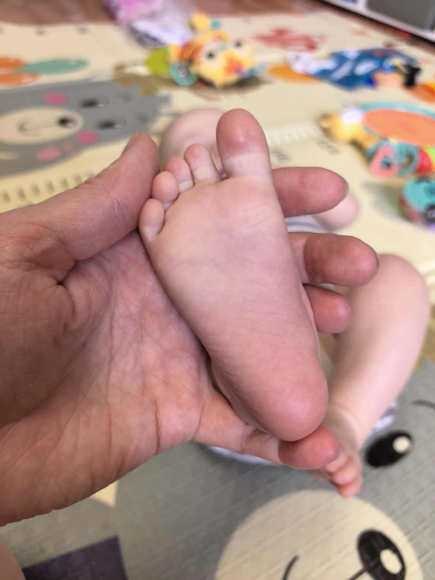 An adult hand holds a baby's foot. The baby is lying on a patterned play mat with toys scattered around. The background is blurred. The location in the city of Фролы, Russia is not possible to ascertain from the given image.