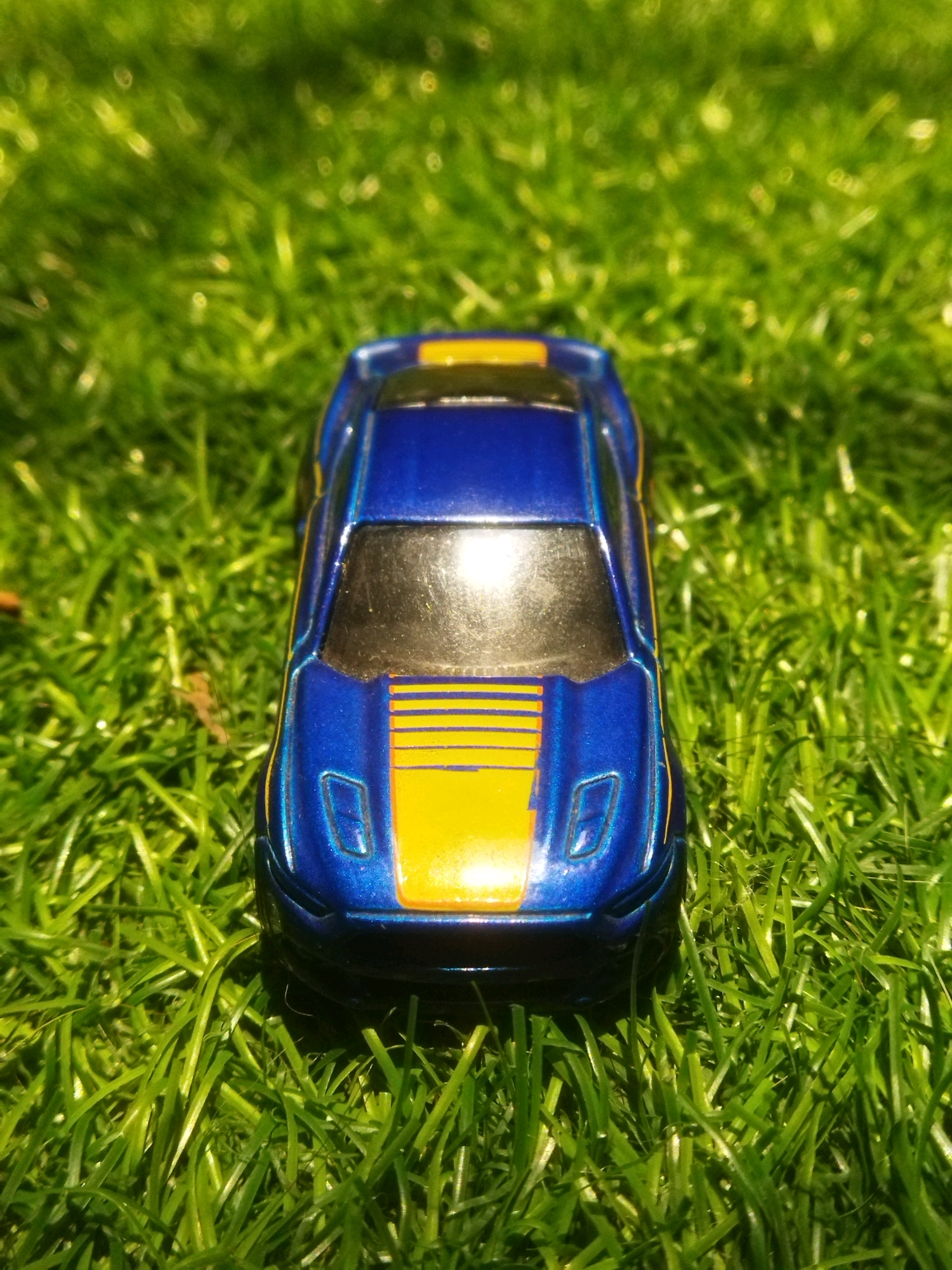 A blue toy car with yellow racing stripes is positioned on a bed of green grass. The car is facing forward, with its hood and windshield prominently visible. The grass appears to be well-maintained and is lit by sunlight, creating a bright and airy atmosphere. The scene is devoid of any human presence or activity. The location is identified as Tarai Bangun, Indonesia.