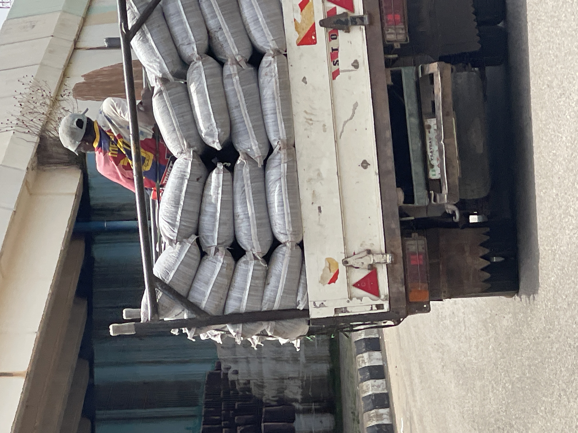 A medium shot depicts the back of a truck overloaded with sacks, parked on a paved road, likely within an urban environment. An individual wearing a helmet, a light-colored top, and a red shirt or jacket is visible towards the front of the truck, seemingly tending to the load. The sacks are densely packed and secured with a metal frame. The back of the truck includes typical truck features such as lights, reflectors, and mud flaps. The location is inferred to be Jos, Nigeria, but lacks definitive contextual clues within the image to confirm the specific location. The surrounding environment consists of architectural structures and greenery, suggesting a street scene.
