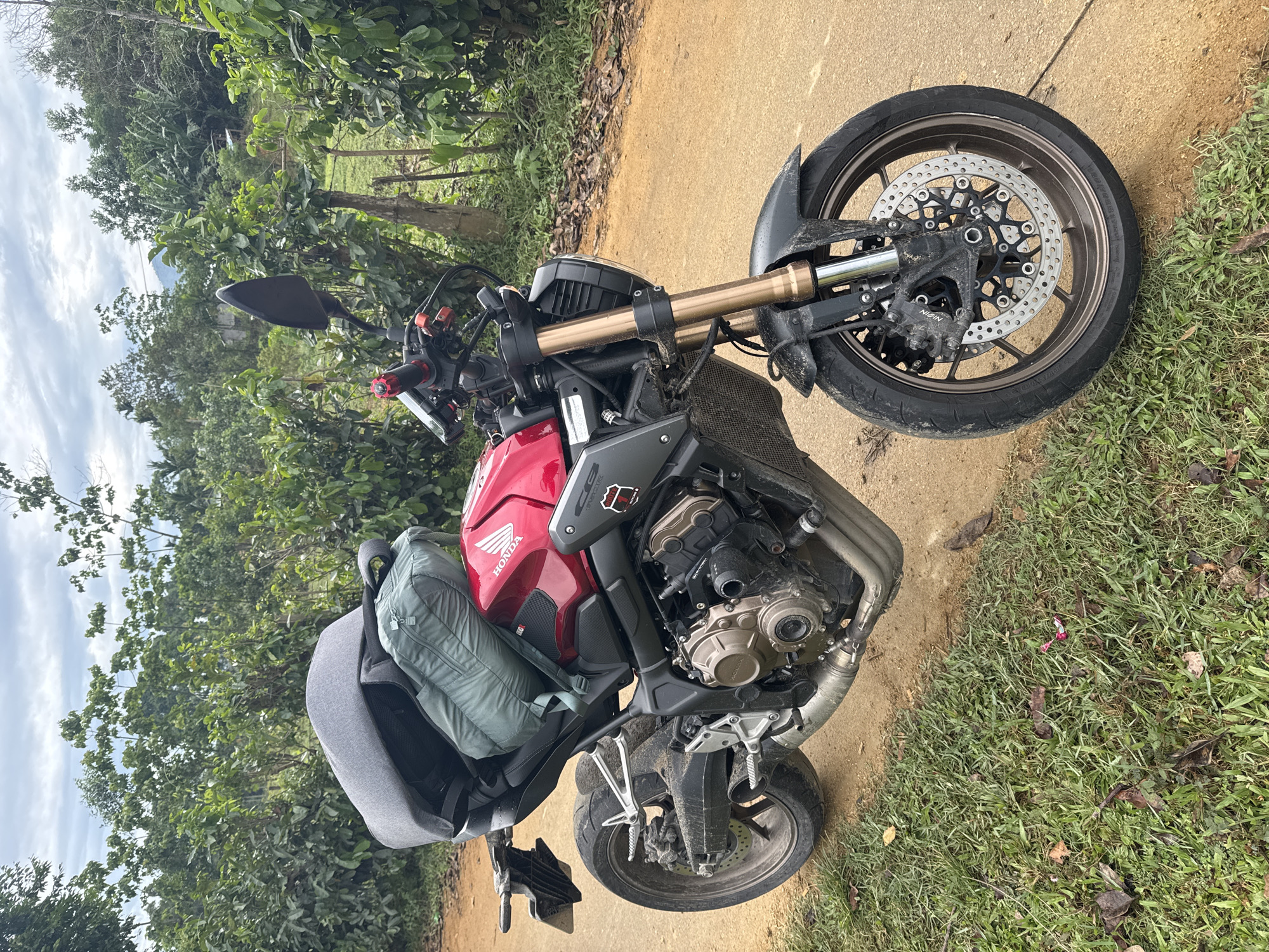 A red Honda motorcycle is parked on a dirt path next to a grassy area, surrounded by lush green vegetation under a cloudy sky. The motorcycle, a naked sportbike, features gold-colored upside-down front forks, dark wheels, and visible engine components. A dark grey backpack is strapped to the rear passenger seat, with a lighter green bag placed on top of it, suggesting luggage for a journey. The front wheel and engine area show some dirt and dust, indicating recent travel on unpaved roads.
Notable details include the "HONDA" logo clearly visible on the red fuel tank and "NISSIN" on the front brake caliper. In the background, beyond the dense foliage, a mountain or large hill can be seen under the overcast sky. The setting suggests a rural or natural environment, likely in the Đà Nẵng, Vietnam area as per the provided context. The time of day is daytime, with soft, diffused lighting due to the cloudy conditions. The motorcycle appears to be stopped, perhaps for a break during a ride.
