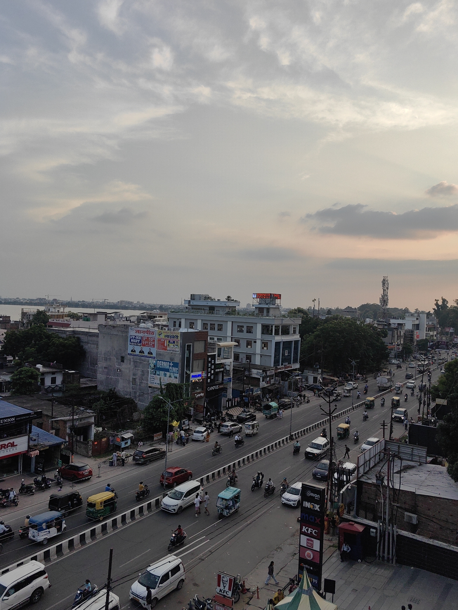 The image depicts a busy street scene in Bichhia, India, likely in the late afternoon or early evening, as indicated by the soft, diffused light and the hues in the sky. The perspective is from an elevated viewpoint, looking down at a multi-lane road filled with traffic.
Vehicles are a dominant feature. Numerous cars, motorcycles, and auto-rickshaws are visible, with some moving and others stopped in traffic. The vehicles vary in color and size, creating a mosaic of movement and activity. Among the vehicles, a yellow auto-rickshaw is prominent in the middle lane, and several white cars are also noticeable. Motorcycles, often with two riders, are a frequent sight.
Along the sides of the road, buildings of varying heights line the street. These include commercial establishments, some with prominent signage. One building displays "KFC" and "INOX," suggesting a commercial complex or mall. Another sign reads "DERMA CLINIC." Further in the distance, taller buildings and structures, including what appears to be a telecommunications tower, are visible. Trees and vegetation are interspersed between the buildings and along the roadside, adding touches of green to the urban landscape.
The sky is partly cloudy, with soft grey and pinkish hues, suggesting a sunset or overcast conditions. The clouds are spread out, with some areas appearing brighter than others.
People are present, though small in scale due to the elevated viewpoint. Individuals are seen walking on sidewalks, riding on motorcycles, and interacting in small groups near the buildings. Their actions are consistent with daily urban life, such as commuting, shopping, or engaging in street-level activities.
The overall impression is one of a dynamic and functional urban environment, characterized by the flow of traffic, the presence of commercial activity, and the everyday lives of its inhabitants within the context of a developing Indian city.