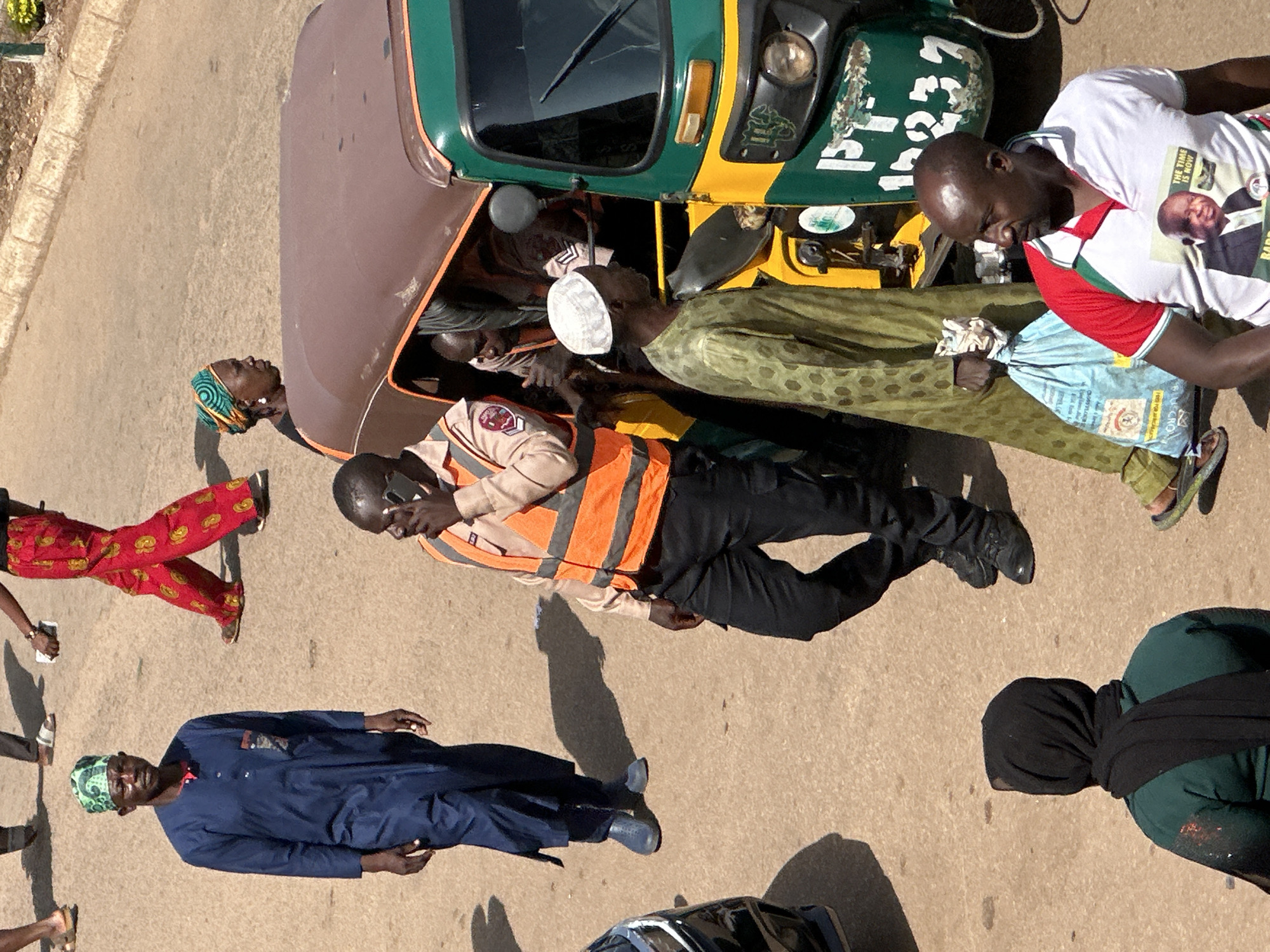 In Bukuru, Nigeria, a street scene unfolds under daylight. A man in a dark blue kaftan and green headscarf stands facing left, his hands at his sides. Behind him, a person in red and yellow patterned trousers walks away, their left hand holding a smartphone. To the right, another individual in a similar vibrant outfit is partially visible.
In the foreground, a man wearing an orange safety vest over a light-colored shirt and black trousers is on the phone, his right hand holding the device to his ear. He stands near a three-wheeled vehicle, a tuk-tuk, which is predominantly green and yellow. Inside the tuk-tuk, a person with a white kufi and a patterned green robe is seen from the side. Other figures are partially obscured within the vehicle.
Further right, a man in a white polo shirt with red sleeves, featuring a printed image of a politician with the text "THE TIME IS NOW," looks to the left, his left arm obscured. He stands next to a light blue shopping bag. In the lower left, a person wearing a dark green outfit and a black hijab is seen from behind, their right arm showing some red and white marks. The ground is paved with a sandy, reddish-brown surface.