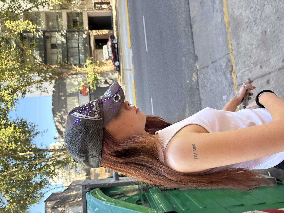 A fair-skinned woman wears a gray bedazzled baseball cap and a white tank top. She has red hair and a nose ring. Her left arm has a small tattoo that reads “self love”. She stands near a green recycling bin. In the background are trees and buildings. On the other side of the road is an empty street with parked cars. The photo is taken during daylight hours with natural lighting.