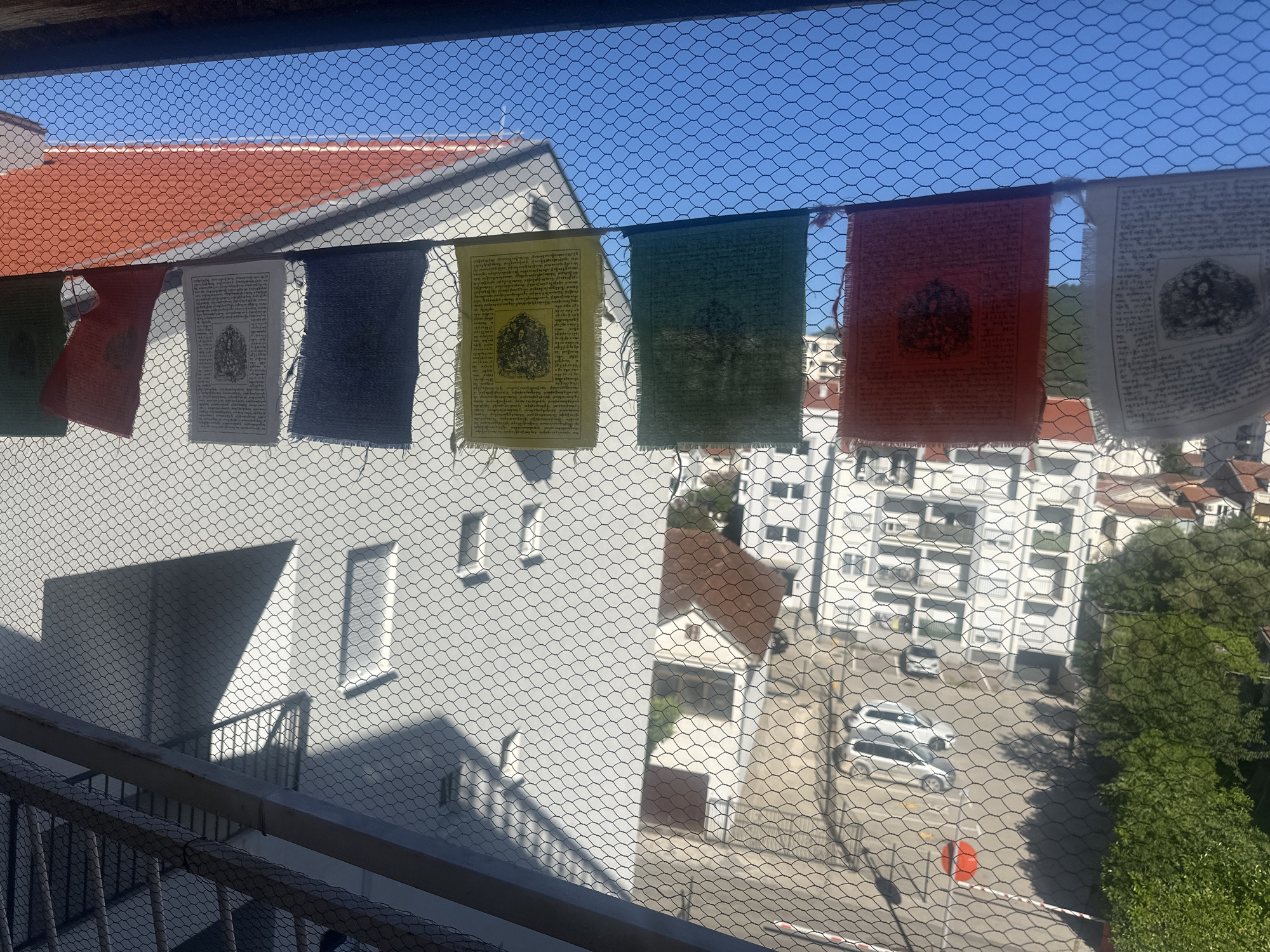 The image shows a view from a balcony or window in Budva, Montenegro. A string of Tibetan prayer flags hangs across the frame. The foreground includes the balcony railing and a wire mesh screen. Through the screen, the view encompasses a white residential building with a red-tiled roof. Further in the distance are other buildings, trees, and a parking area with several cars. The sky is visible above the buildings and is clear and blue.