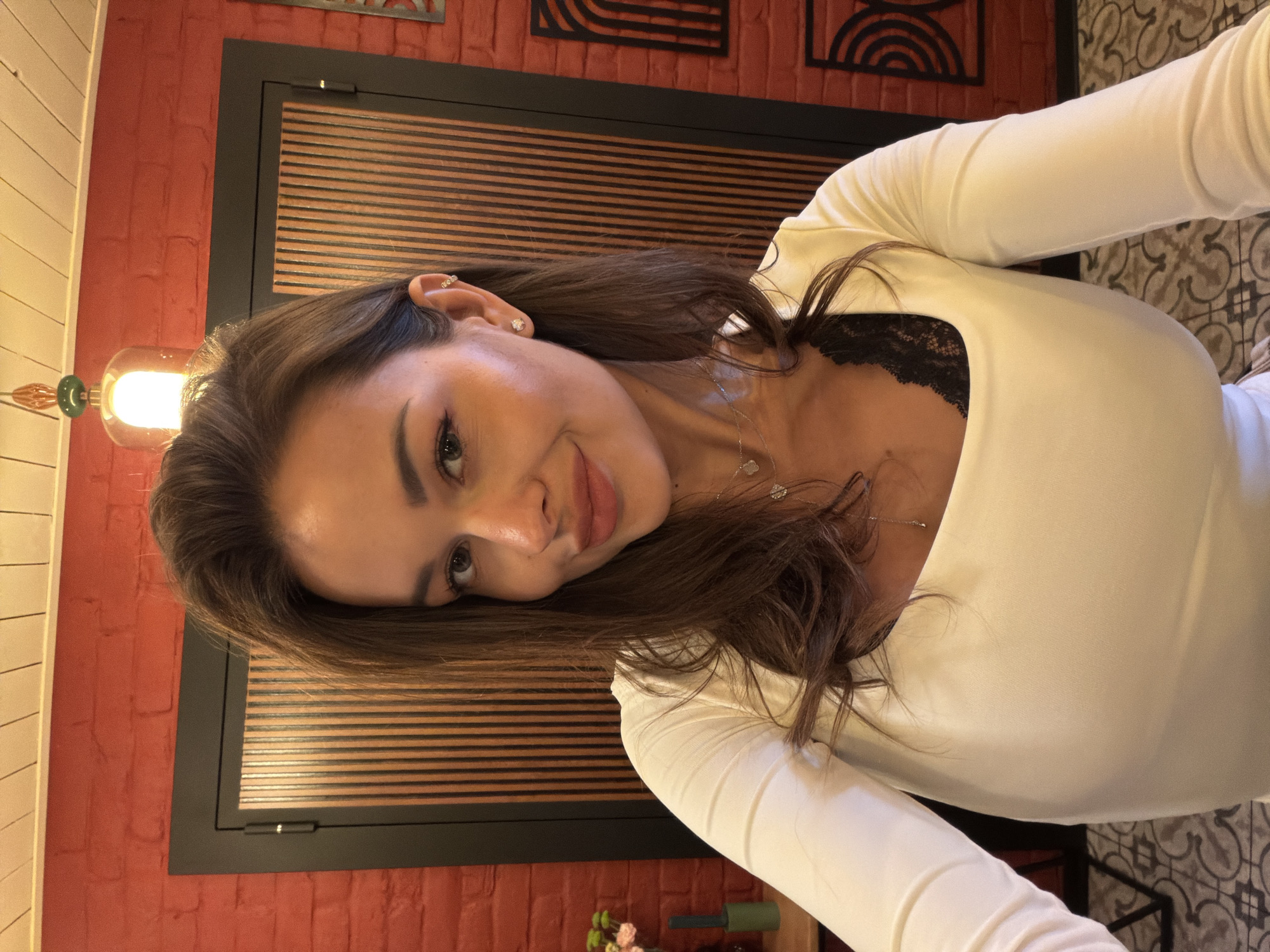 A woman with long, dark brown hair and fair skin takes a selfie in what appears to be a dimly lit restaurant or cafe. She is wearing a white, long-sleeved shirt with a square neckline, revealing a glimpse of black lace underneath. She has on delicate jewelry, including a stud earring in her right ear and a two-layered necklace with small pendants. Her makeup is subtle, with a focus on her eyes and lips. The background features a red brick wall, a dark wooden partition with vertical slats, and a decorative light fixture with a green accent at the top. A small vase with pink and white flowers is visible on the left. The floor has an intricate tiled pattern. The overall impression is of a casual, indoor setting.