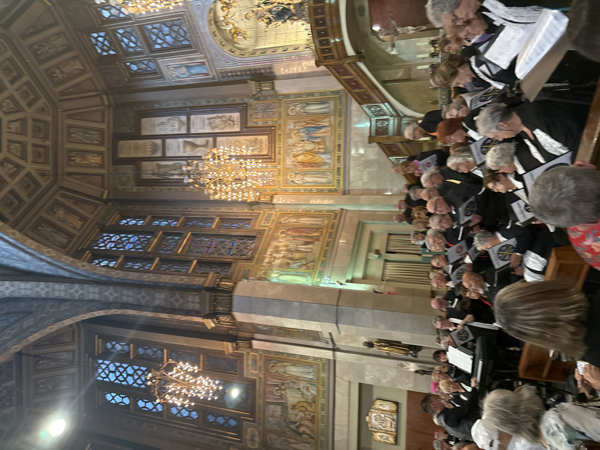 This image captures a choir performing inside a grand cathedral, likely in Barcelona, Spain, given the architectural style. The setting is ornate, with high, vaulted ceilings adorned with decorative paintings and intricate stonework. Large, stained-glass windows with geometric patterns let in diffused light. Chandeliers with numerous lights hang from the ceiling, illuminating the space.
In the foreground, a choir of approximately 30-40 individuals, of varying ages, are arranged in rows, holding music sheets. They are dressed in black choir robes with white accents. Some members are looking at their music, while others are looking forward, seemingly at a conductor or towards an unseen focal point. A man in a black shirt and vest stands to the left, appearing to direct the choir, possibly playing a keyboard. In front of the choir, a few audience members are visible, seated on pews.
The background reveals further architectural details of the cathedral, including detailed murals on the walls depicting religious scenes and figures. A wooden staircase with a railing ascends to a higher level on the right side of the frame. A statue of a religious figure is visible on this upper level. The overall atmosphere is solemn and focused on the performance within the majestic religious structure.