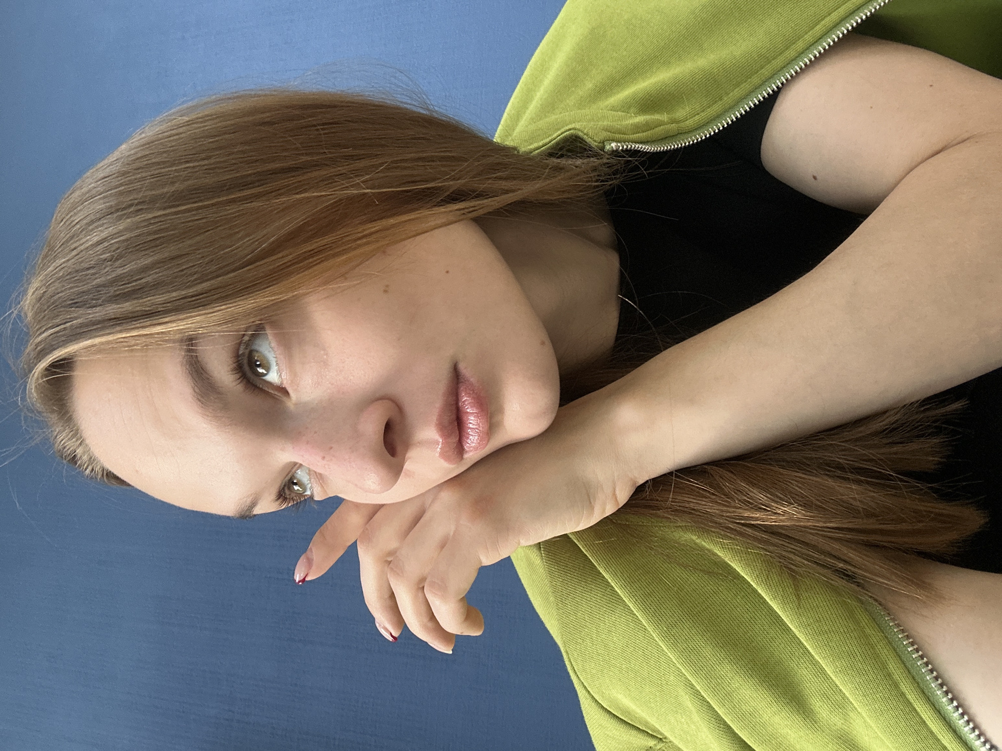 In the image, a young woman with long, light brown hair is pictured against a plain blue background. She is wearing a bright green zip-up jacket over a black shirt. Her right hand is raised to her face, with her fingers lightly touching her cheek and temple, and her gaze is directed upwards and to the left. The lighting suggests an indoor setting. The image does not provide any specific location information within Chaikovsky, Russia.