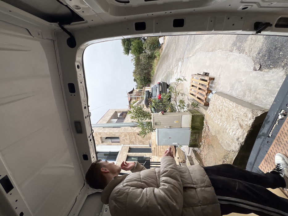 The image is taken from within the white-painted cargo area of a vehicle, looking out onto a street in Bigova, Montenegro, under an overcast sky.
In the foreground, on the left, a male subject stands inside the vehicle, facing away from the camera and looking towards the exterior. He wears a light beige puffer jacket, dark track pants with three white stripes, and white sneakers. His right hand is raised towards his mouth, and his left hand holds a small white object.
The scene outside the vehicle shows a paved street, wet from recent precipitation, sloping gently uphill. To the left, a modern, light-colored multi-story building with dark balcony railings is visible. In front of this building, two grey utility cabinets are situated on a concrete base; one displays a red lightning bolt symbol. To the right of the cabinets, a dark grey hatchback vehicle with a roof rack and black cargo box is parked facing away from the camera. A wooden shipping pallet lies on the wet ground near the edge of the street, with plastic bottles visible beneath it. Green foliage and bushes line the side of the road further down the street. The ground directly outside the van consists of broken concrete and wet dirt.
