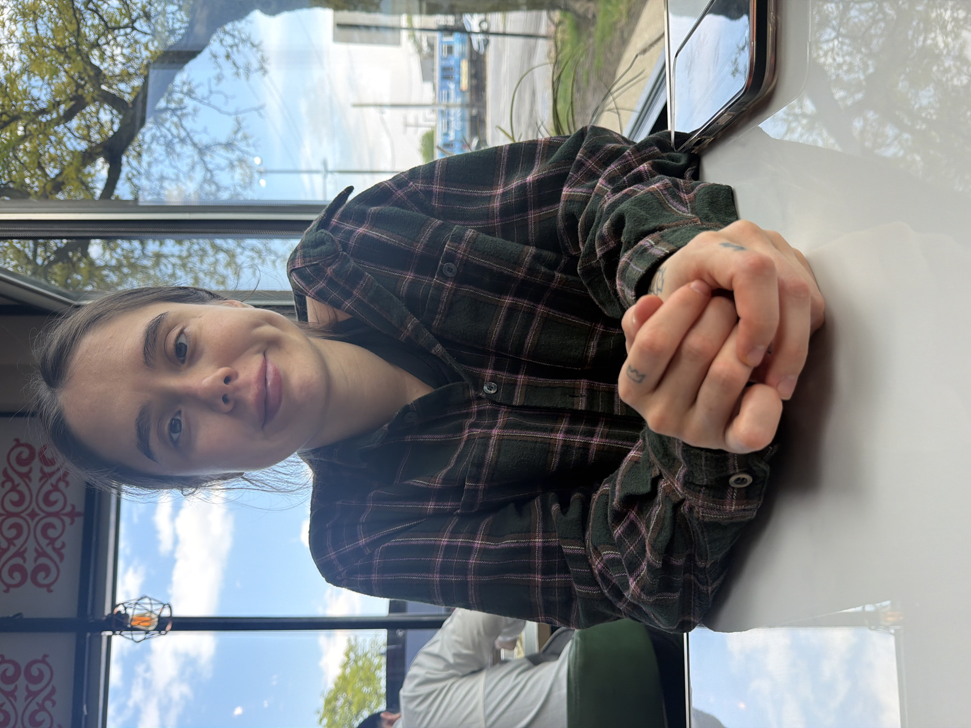 A medium shot depicts a young woman, likely in her early to mid-20s, seated at a table indoors. She is fair-skinned with dark hair pulled back from her face. She is wearing a dark, patterned flannel shirt and her hands are clasped together on the table. The background reveals a daytime urban scene visible through a large window. Trees are visible outside, along with the architectural details of buildings that suggest a cityscape. Reflections in the window, particularly the sky and clouds, add to the impression of an outdoor setting. The restaurant appears to be located in Chicago.