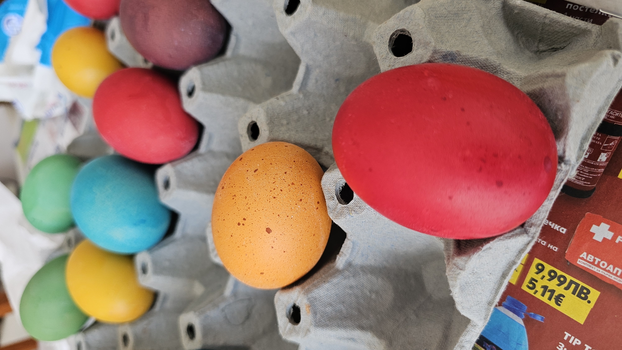 The image shows a close-up of colorful Easter eggs in a gray cardboard egg carton. The eggs are dyed in various colors including red, orange, purple, blue, green, and yellow. Some of the eggs have slight blemishes or spots on their surfaces. The carton is placed on top of a magazine or advertisement with Bulgarian text and pricing in both Bulgarian Lev (лв) and Euros (€). The text on the magazine includes "автоаптечка" and "комплект за първа медицинска помощ," suggesting a first aid kit advertisement. The scene appears to be indoors, possibly a home setting in Bulgaria. There is no explicit mention or hint of Varna, Bulgaria in the image.