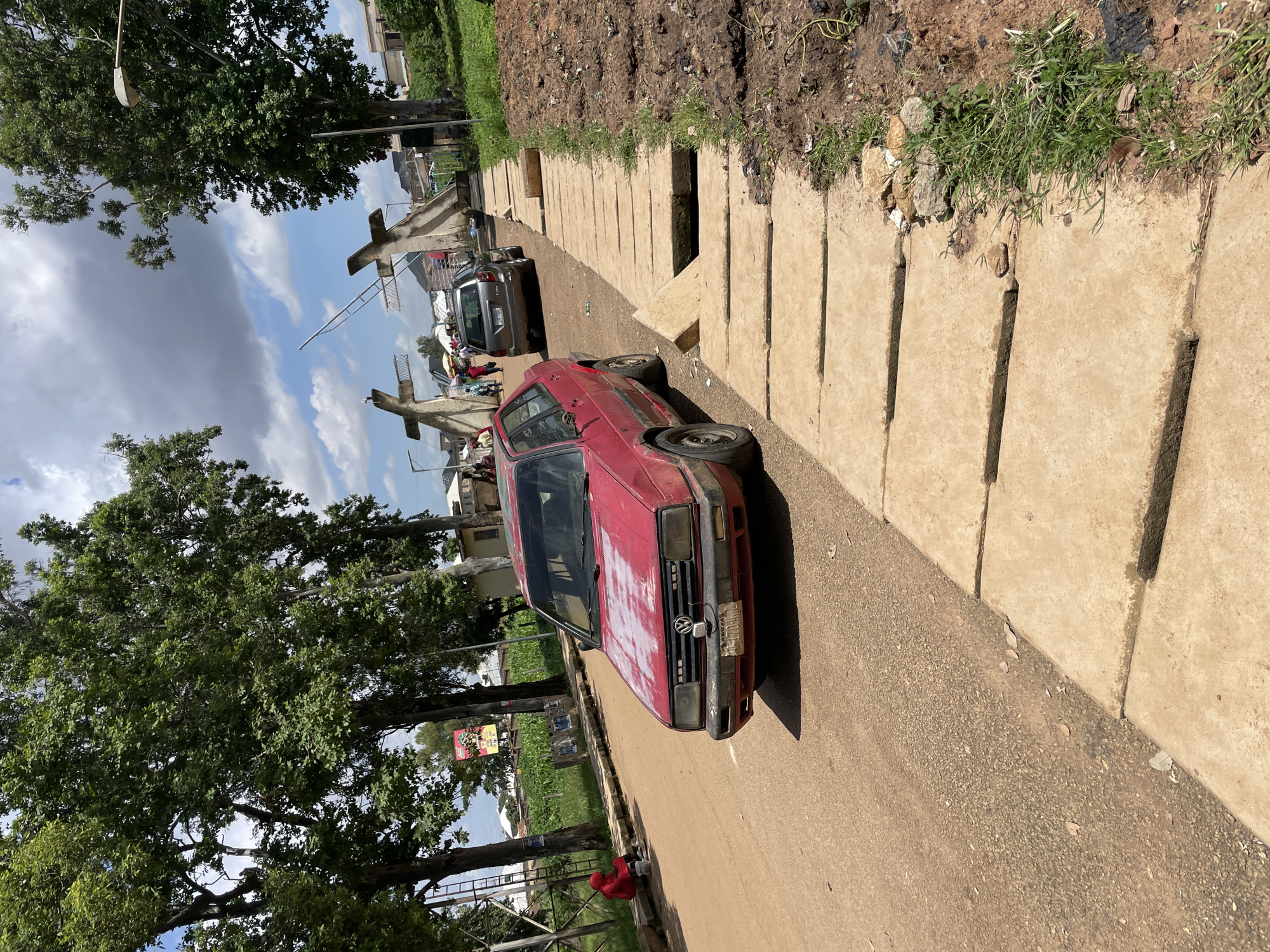 The image depicts a street scene in Rafin Par, Nigeria. In the foreground, a red Volkswagen car is parked on a dirt road. To the right of the car, a concrete drainage system with large rectangular slabs runs along the side of the road. Some of the slabs are broken or displaced, revealing a gap underneath. Adjacent to the drainage, a patch of rough ground with sparse green grass and dirt is visible.
In the background, a silver SUV is also parked on the road, further down and approaching an entrance structure. This structure appears to be a gateway or archway with two large, stylized cross-like concrete pillars. People are visible walking through this gateway. Tall trees with lush green foliage line the left side of the road, with some buildings and what appears to be agricultural fields visible beyond them. A person wearing a red hoodie is seated on the curb to the left, near a white bucket. The sky above is partly cloudy.