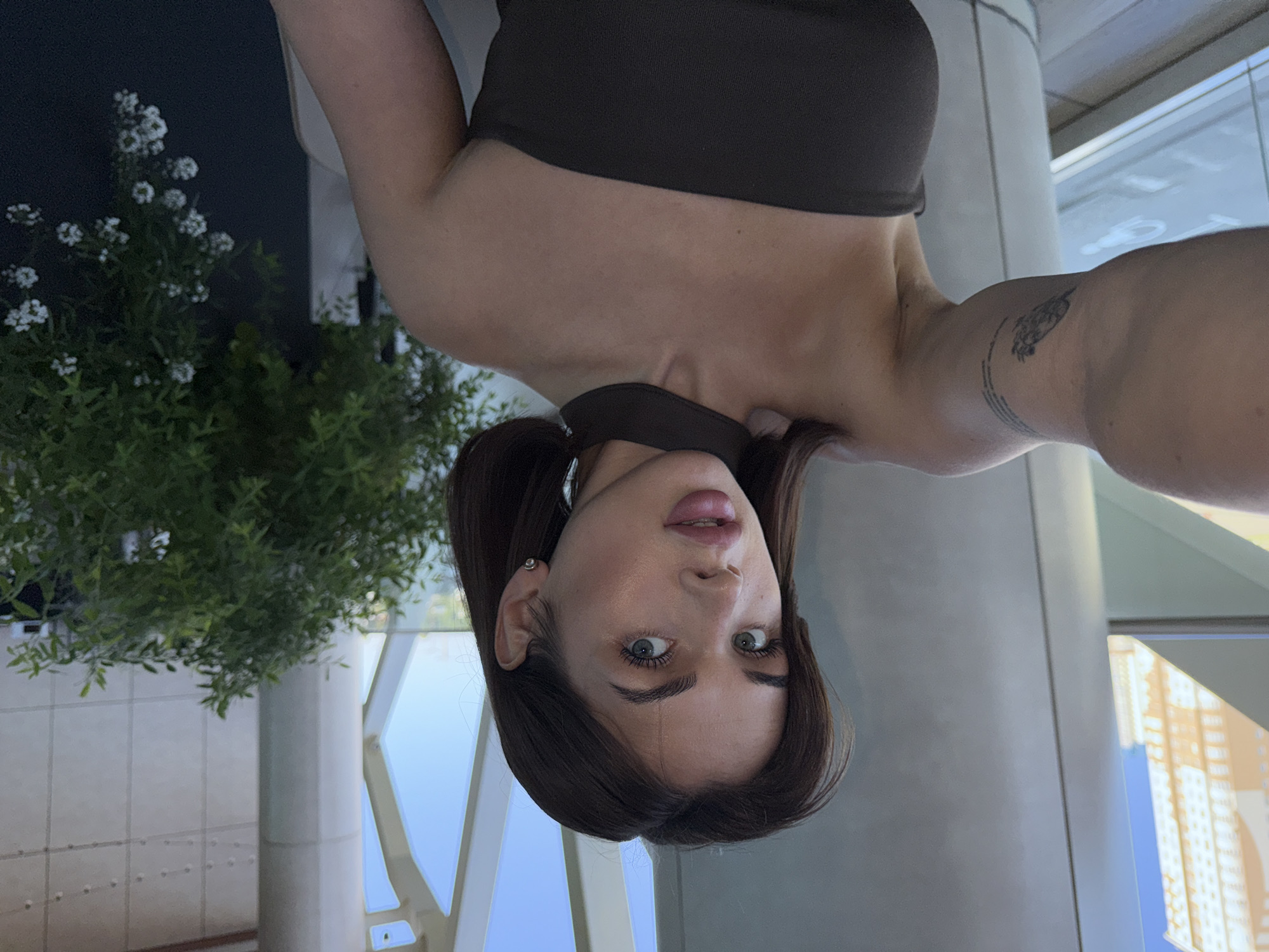 A young woman with brown hair and blue eyes is taking a selfie in what appears to be an indoor, modern building with large windows. She is wearing a dark, strapless top and a choker. Her left arm is extended towards the camera, revealing a tattoo on her upper arm. Behind her, there are large pillars, a potted plant with white flowers, and a view of a city skyline with tall buildings, suggesting an urban setting. The light suggests it is daytime. The specific location in the city of Perm, Russia, is not identifiable from the image alone.