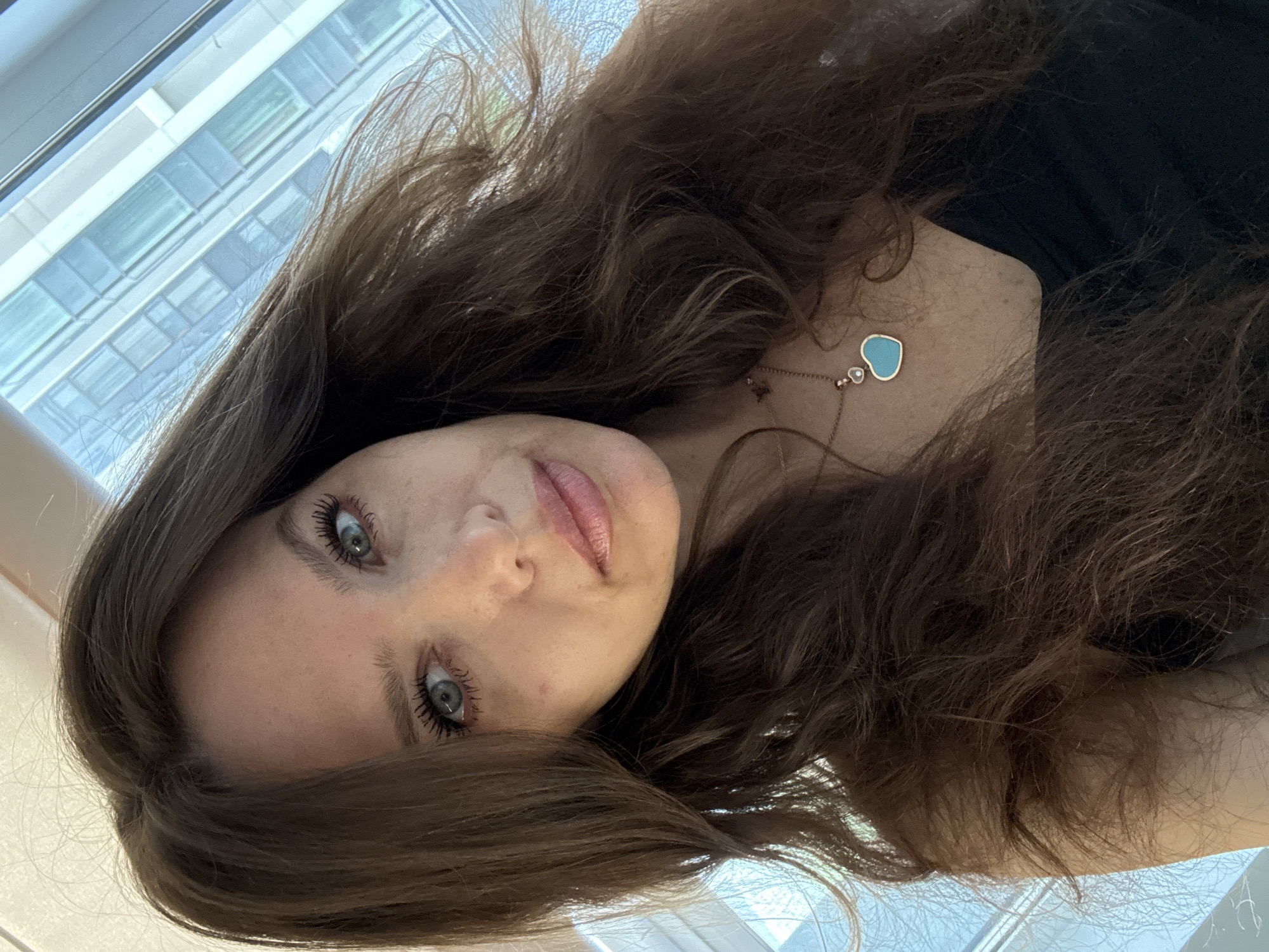 A young woman with long, wavy brown hair is positioned in the foreground. She has fair skin, blue eyes, and is wearing mascara. A blue heart-shaped pendant hangs from a delicate rose-gold necklace around her neck. She is wearing a black top. The background shows a window with a view of a modern building with multiple windows. The location is Khimki, Russia, based on the architectural style of the building visible through the window.