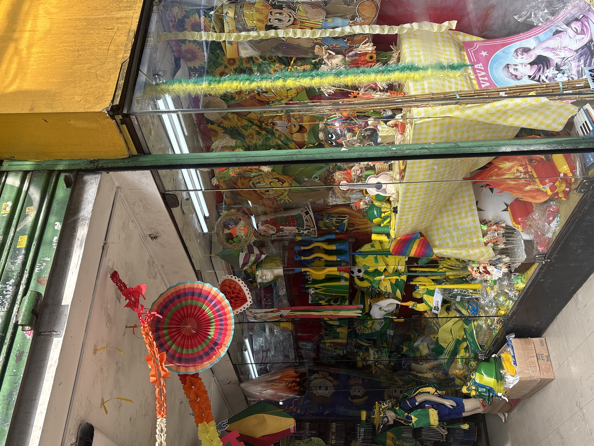 This image shows the storefront of a shop in São Paulo, Brazil, filled with festive merchandise, likely for a holiday or event. The shop's facade is painted yellow, with a green metal shutter partially rolled up above the entrance. The display window is packed with various items, predominantly in yellow and green, suggesting a connection to Brazilian national colors or a specific celebration.
In the foreground, a mannequin dressed in blue shorts, a yellow vest, and a matching hat with a green scarf stands on a cardboard box. The window itself is filled with an array of merchandise. Hanging decorations, including colorful paper garlands and a large circular fan-shaped ornament, are visible at the top. The interior is stocked with items such as hats, flags, noisemakers, plastic toys, and effigies of straw figures. A mannequin head wearing a green and yellow hat is also displayed.
A table covered with a yellow and white checkered tablecloth is visible inside, adorned with more merchandise, including a guitar, straw figures, and small toys. In the lower right corner, a poster with the word "VIVA" and an image of two figures can be seen. The overall impression is a vibrant and cluttered shop display, characteristic of a street vendor or small business catering to celebratory occasions in São Paulo. The environment appears to be an urban setting, with the shop front opening onto a street or sidewalk.