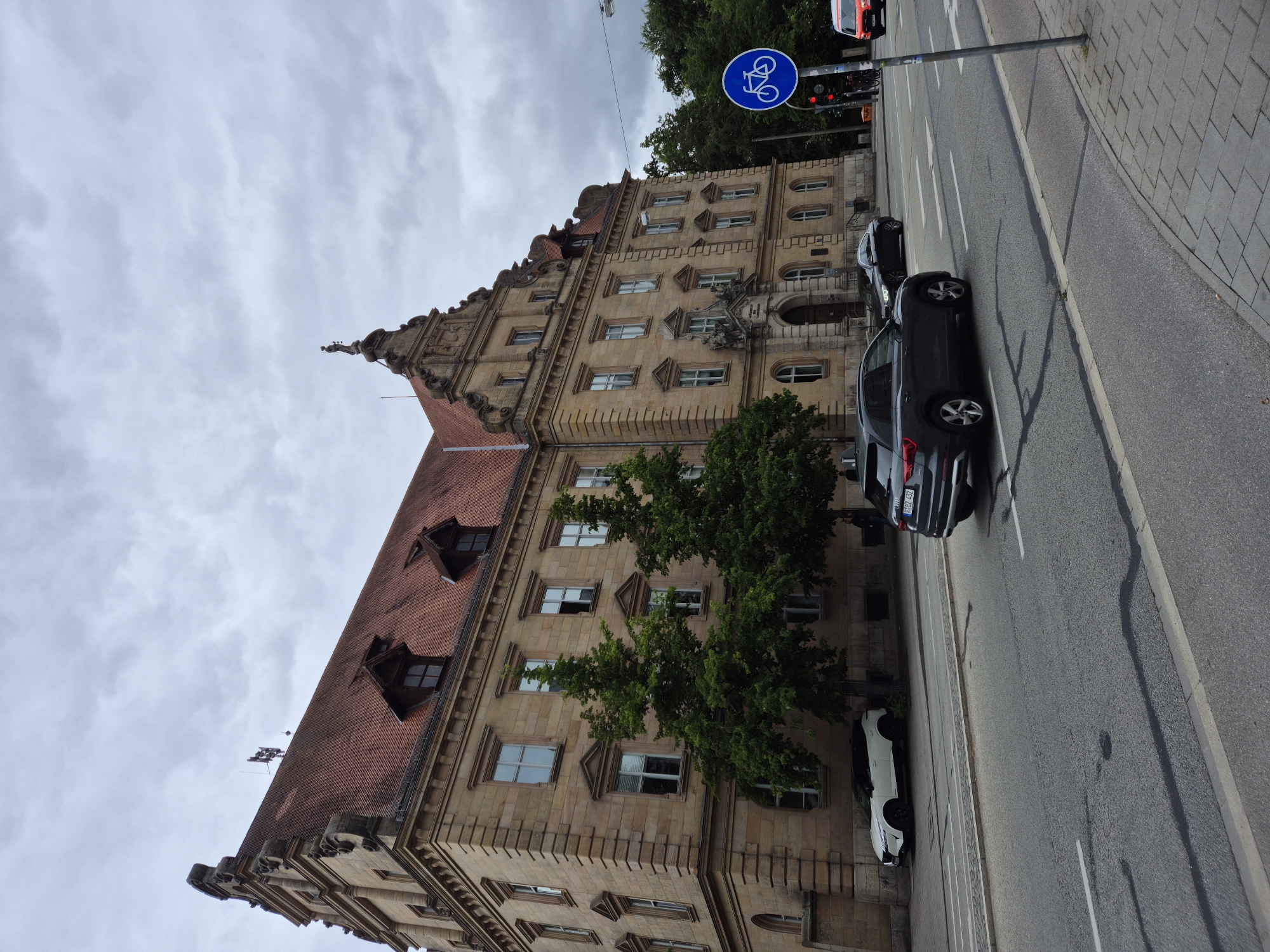 The image depicts the Landgericht Regensburg, a courthouse located in Regensburg, Germany. The building is a grand, multi-story structure with a red tile roof, adorned with decorative architectural elements and several dormer windows. In the foreground, a street runs alongside the building, with cars visible on the road. A dark grey SUV is in the immediate foreground, with a white SUV parked on the side of the street further down. A blue circular sign with a white bicycle symbol is mounted on a pole on the right side of the image, indicating a bicycle lane. Traffic lights are also present. Green trees line the street, partially obscuring the lower facade of the building. The sky is overcast with clouds, suggesting a cloudy day.