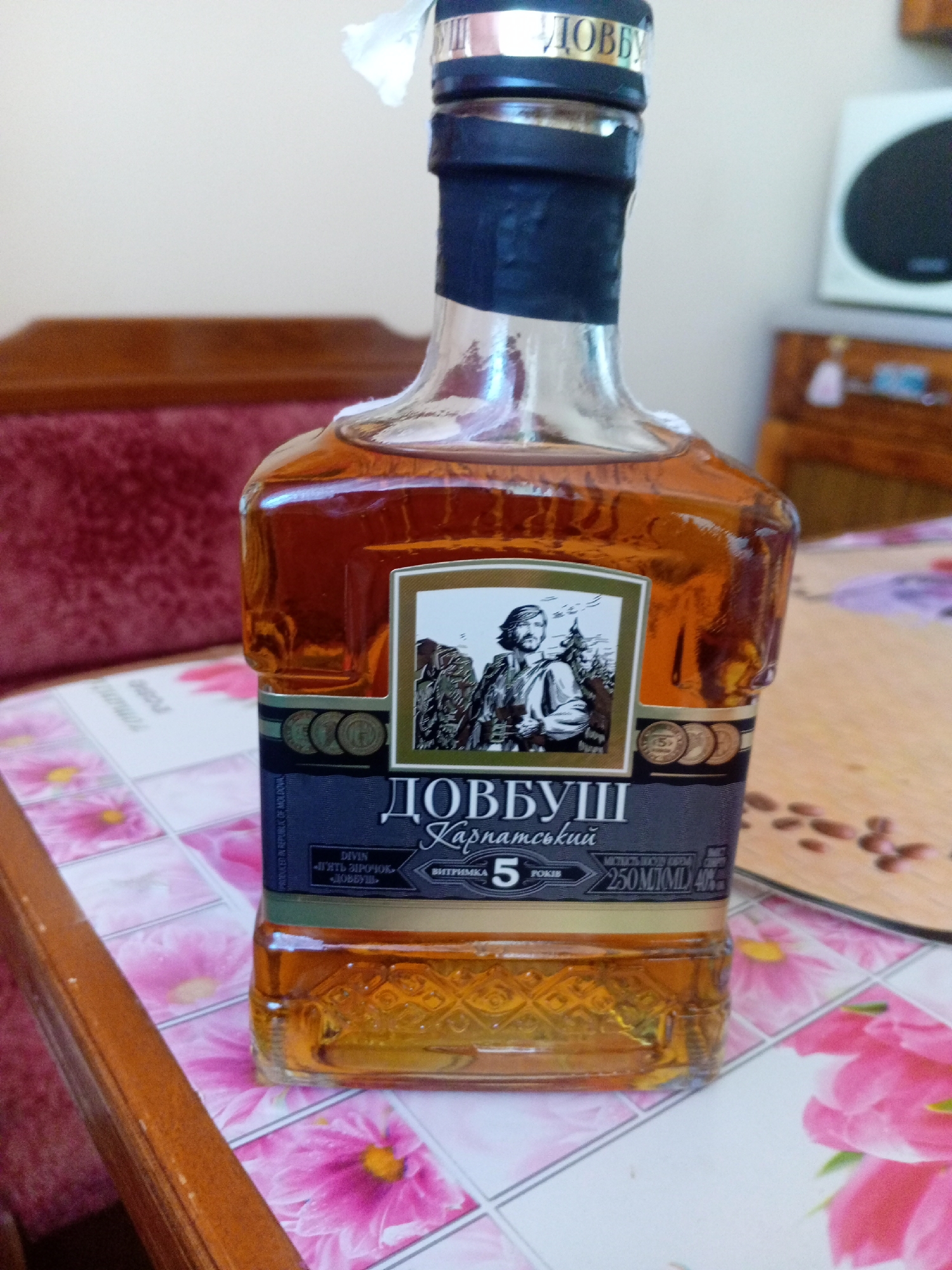 A close-up, eye-level, vertical, indoor shot captures a bottle of Dobbush Carpathian Cognac. The bottle, made of clear glass, contains a golden-amber liquid. A dark blue label wraps around the lower part of the bottle, featuring a portrait in a square inset. The portrait depicts a man in traditional clothing with a beard, standing in a mountainous landscape. The label also displays text in Cyrillic script, including "ДОВБУШ" and "Карпатський," as well as numerical and alphabetical indicators for volume and alcohol percentage. The bottle rests on a table covered with a pink and white floral patterned tablecloth. To the right of the bottle, a wooden cutting board with scattered small brown objects, possibly coffee beans, is visible. In the background, a wooden piece of furniture and a white speaker with a black circular grille are discernible. The lighting appears to be natural, coming from the left. The scene is likely within a private residence, not a public space in Tiachiv, Ukraine.
