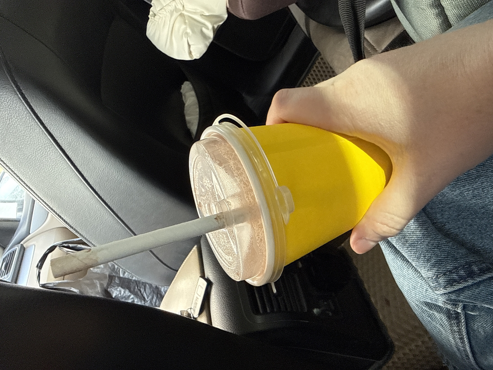 A person's hand holds a bright yellow cup with a clear plastic lid and a white straw inside a car. The hand, illuminated by bright sunlight, casts a shadow on itself. The person wears blue denim jeans, visible in the lower right portion of the frame. The cup contains a light brown or pinkish liquid with a textured surface, possibly from foam or bubbles.
The setting is the interior of a vehicle, likely during daytime given the strong sunlight. Visible car elements include dark, textured upholstery on the left and foreground, a grey woven fabric seat and a dark grey floor mat in the background. A light tan dashboard panel and a dark air vent are also visible through openings in the car's interior. No discernible text is present in the image.