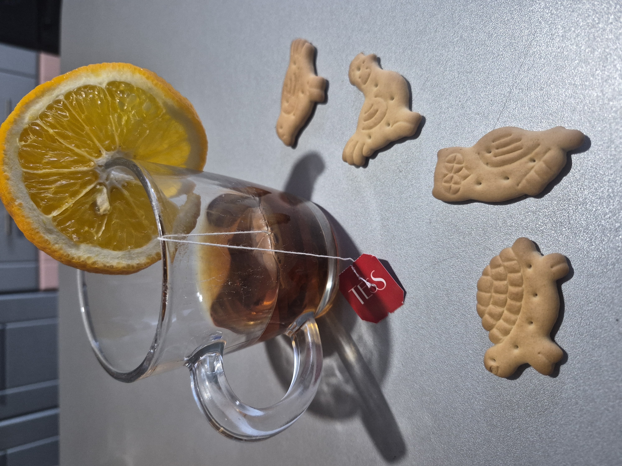 A tilted glass mug containing tea with a tea bag labeled "TESS" sits precariously, adorned with a slice of orange. Scattered nearby are four animal-shaped biscuits. The backdrop is a plain grey surface. The location is unknown.