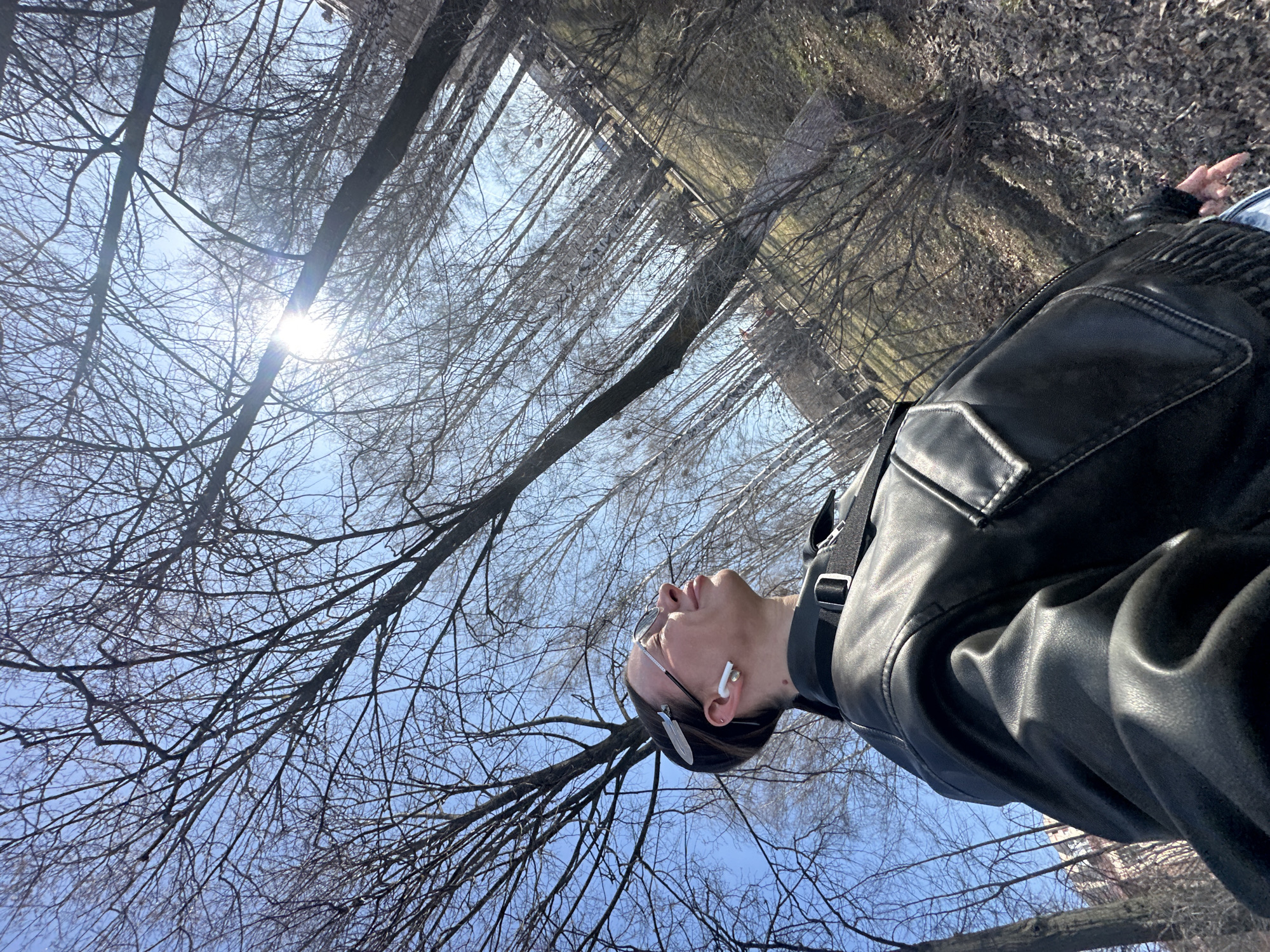 This media file captures an outdoor scene from a low-angle perspective, looking upwards towards a bright sky and tree canopy in Votkinsk, Russia. The primary subject is a person, likely male, positioned on the right side of the frame, viewed from the chest up in a three-quarter profile. The person is wearing a black leather jacket, glasses resting on their head, and white wireless earbuds in both ears. Their head is tilted back, and they are looking directly upwards towards the sky.
The background is dominated by a clear, vibrant blue sky filled with a dense network of bare tree branches, indicating either late autumn, winter, or early spring. The sun is visible in the upper left, casting a strong lens flare and bright light through the branches. Several tree trunks extend vertically and diagonally throughout the composition. The ground, partially visible in the lower right, is covered with dry, fallen leaves and twigs. Distant, indistinct structures or buildings can be faintly observed through the trees.
The activity taking place is the person looking up, seemingly pausing to appreciate the sunlight or the natural environment. The time of day appears to be daytime, given the bright sun and clear sky. The weather is sunny and clear. There is no visible text in the image. The bare trees and ground cover align with the specified location context of Votkinsk, Russia, during colder seasons. The overall mood appears calm and contemplative.