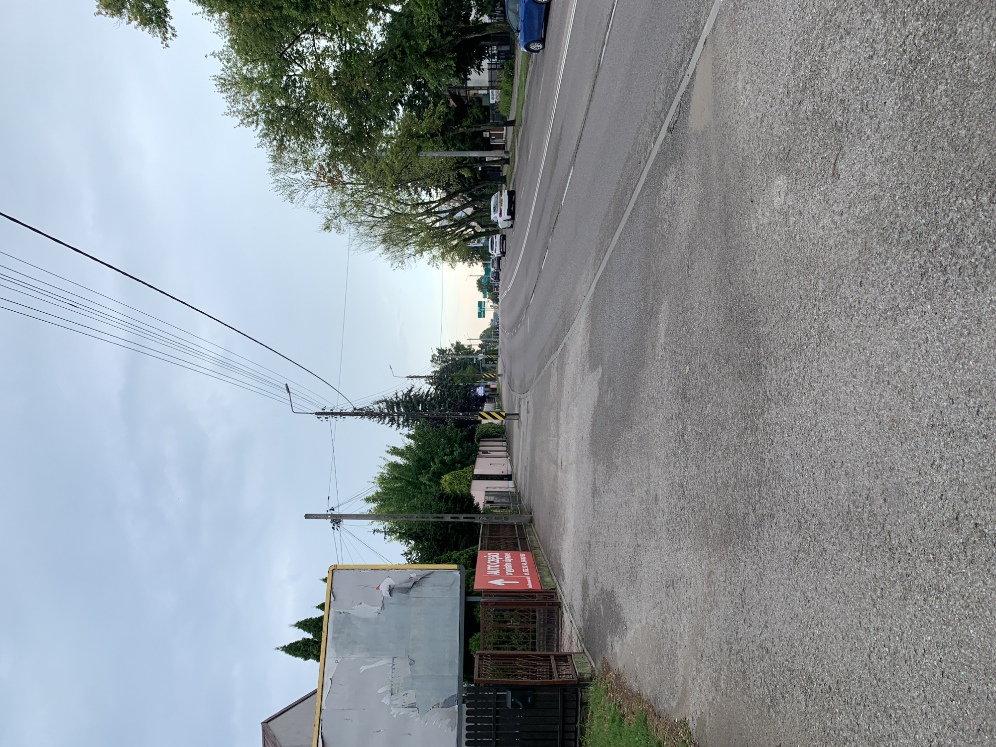 The image depicts a street scene in Białystok, Poland, with a focus on a sidewalk and a road. On the left side of the image, a gray billboard with peeling posters is mounted on a yellow frame, partially obscuring a house with a brown roof. Below the billboard, a black metal fence encloses a property. A red sign with white text and an arrow is attached to the fence, advertising "AUTO CZĘŚCI oryginalne używane" (original used car parts) and a phone number. Further down the street, a row of houses with light-colored facades is visible. The road is asphalt with white lane markings. Several cars are driving on the road, including a white sedan and a blue hatchback. Tall green trees line the right side of the road, with some houses visible behind them. Overhead, power lines crisscross the sky, which is overcast. The overall scene suggests a commercial or suburban area.