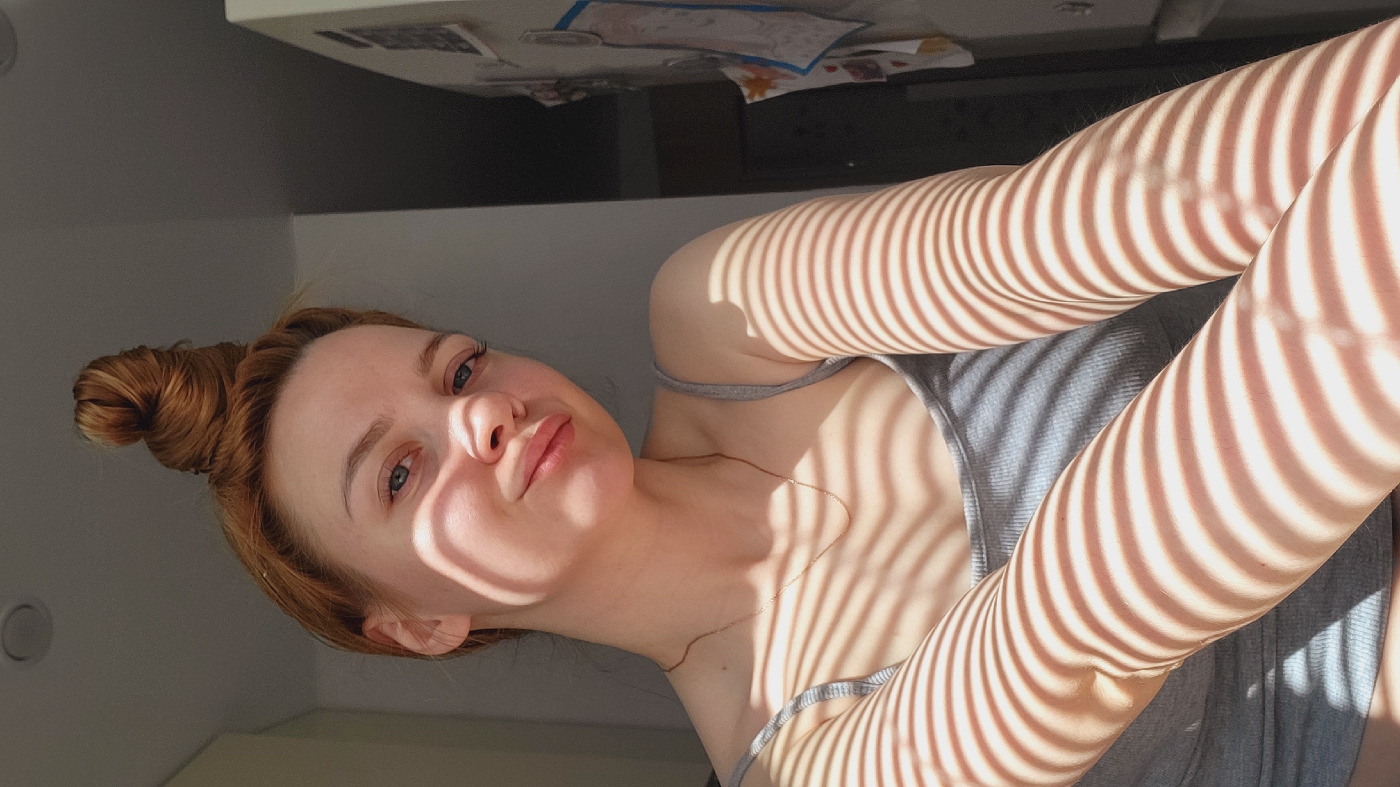 An upward-facing portrait captures a young woman in an indoor setting in Chaikovsky, Russia. Her reddish-brown hair is gathered into a casual bun atop her head. She has light-colored eyes and a fair complexion, with a subtle pout on her lips, looking directly into the camera. She appears to be wearing a simple grey tank top or camisole.
The scene is dramatically illuminated by sunlight filtering through blinds or a similar patterned screen, casting prominent, parallel striped shadows across her face, neck, and arms. These shadows create a striking visual effect, highlighting parts of her skin and clothing while obscuring others. The bright sunlight suggests it is daytime, possibly morning or afternoon.
The background is minimally visible but indicates an interior space with a light-colored ceiling. Above her, a few papers or objects are scattered on what appears to be a desk or shelf. The overall impression is a casual, artistic moment captured with natural light. No discernible text is visible in the image.