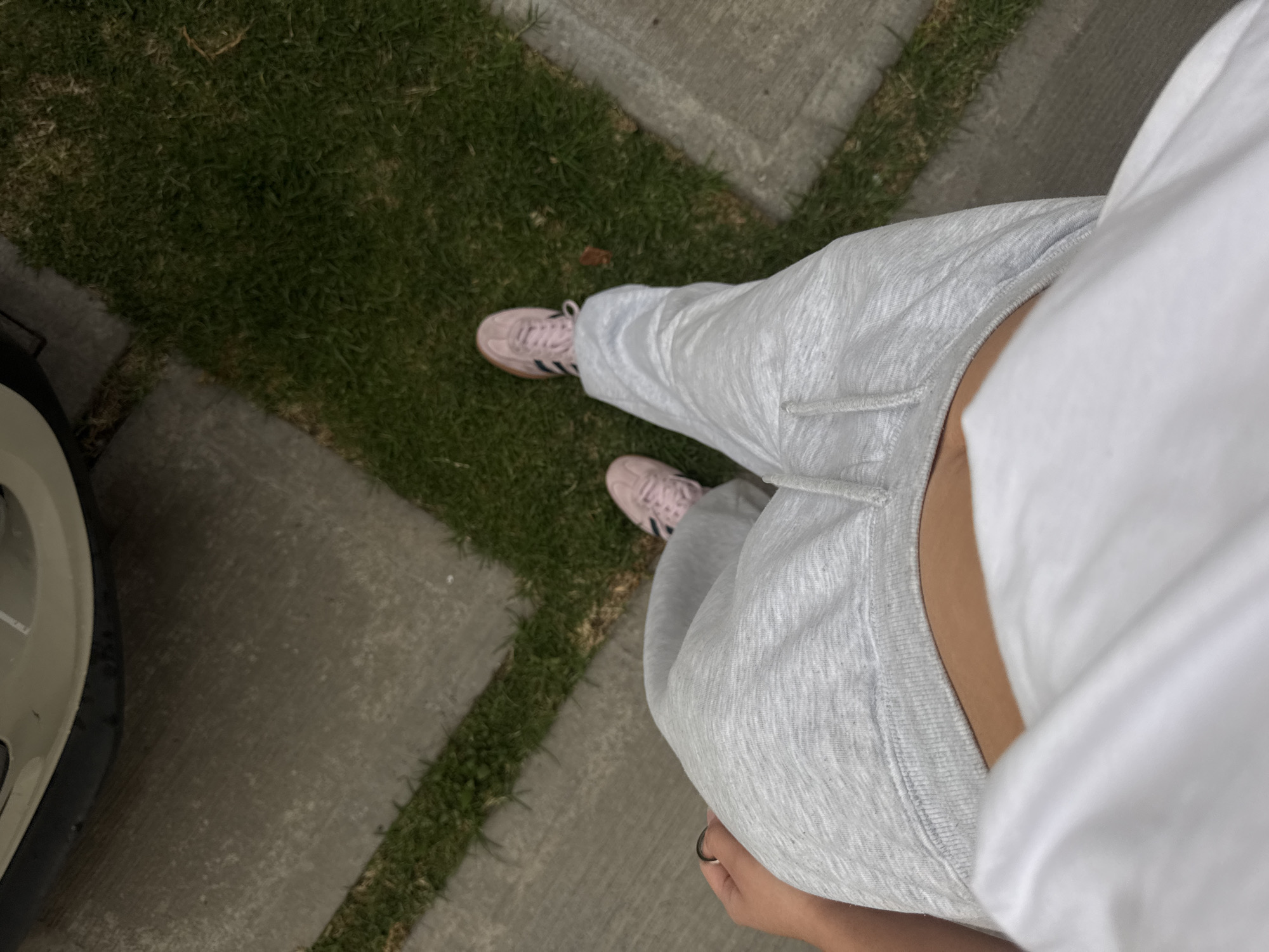 A person wearing light gray sweatpants and pink sneakers stands on a concrete and grass walkway. The corner of a light-colored car is visible to the left. The scene is in an outdoor setting with natural light. No specific location is discernible from the image.