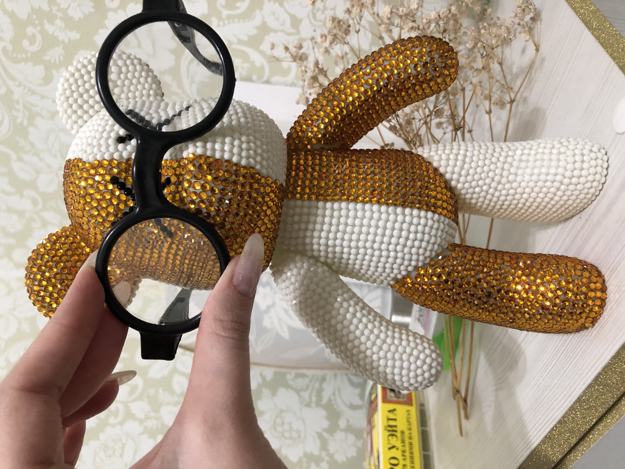 A medium, eye-level, indoor shot depicts a stuffed toy bear partially covered in gold and white rhinestones with a hand holding a pair of black-rimmed clip-on glasses in front of its face.
The bear, positioned on a surface, has its head mostly covered in white rhinestones except for a band of gold rhinestones around its neck. Its body and limbs are similarly decorated with alternating bands of white and gold rhinestones. The bear is lying on its back, with its arms and legs spread out.
A hand with long, painted nails holds a pair of clip-on glasses in front of the bear's face. The glasses are black with round lenses.
Behind the bear, there is a small bunch of dried flowers. A box labeled "PO YZITA" in Russian is visible in the lower portion of the photo.
The surface the bear is on appears to be white with a textured pattern. A golden decorative edge is partially visible at the edge of the surface.
I do not have the information about the location in the city of Noviy, Russia.
