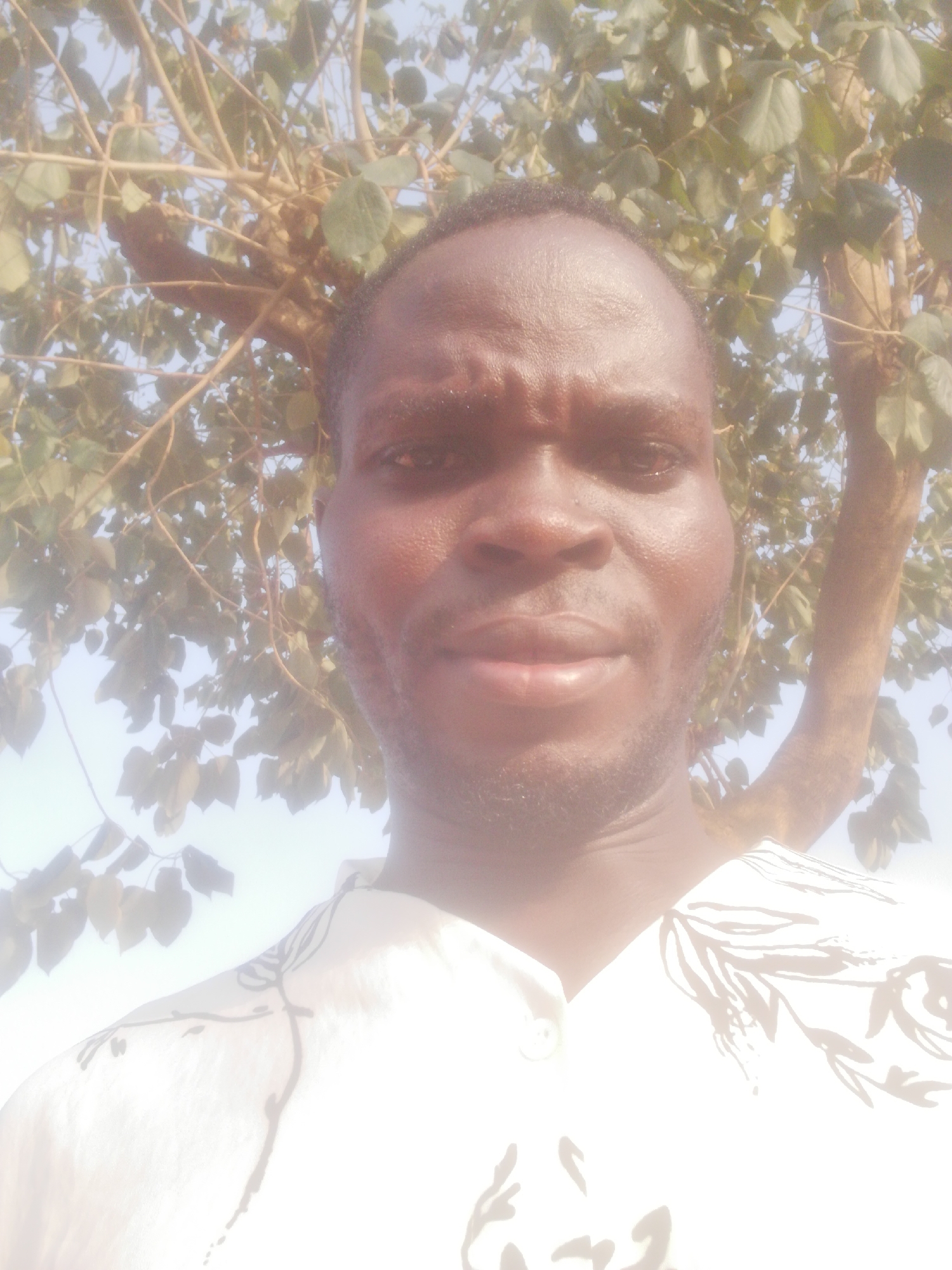 A close-up, slightly low-angle shot features a dark-skinned man standing outdoors. He is facing the camera. He has short hair and a light beard. He is wearing a white patterned shirt. Behind him is a tree with branches and leaves, which is slightly out of focus. The sky is visible in the background. The location is Unknown, Unknown.
