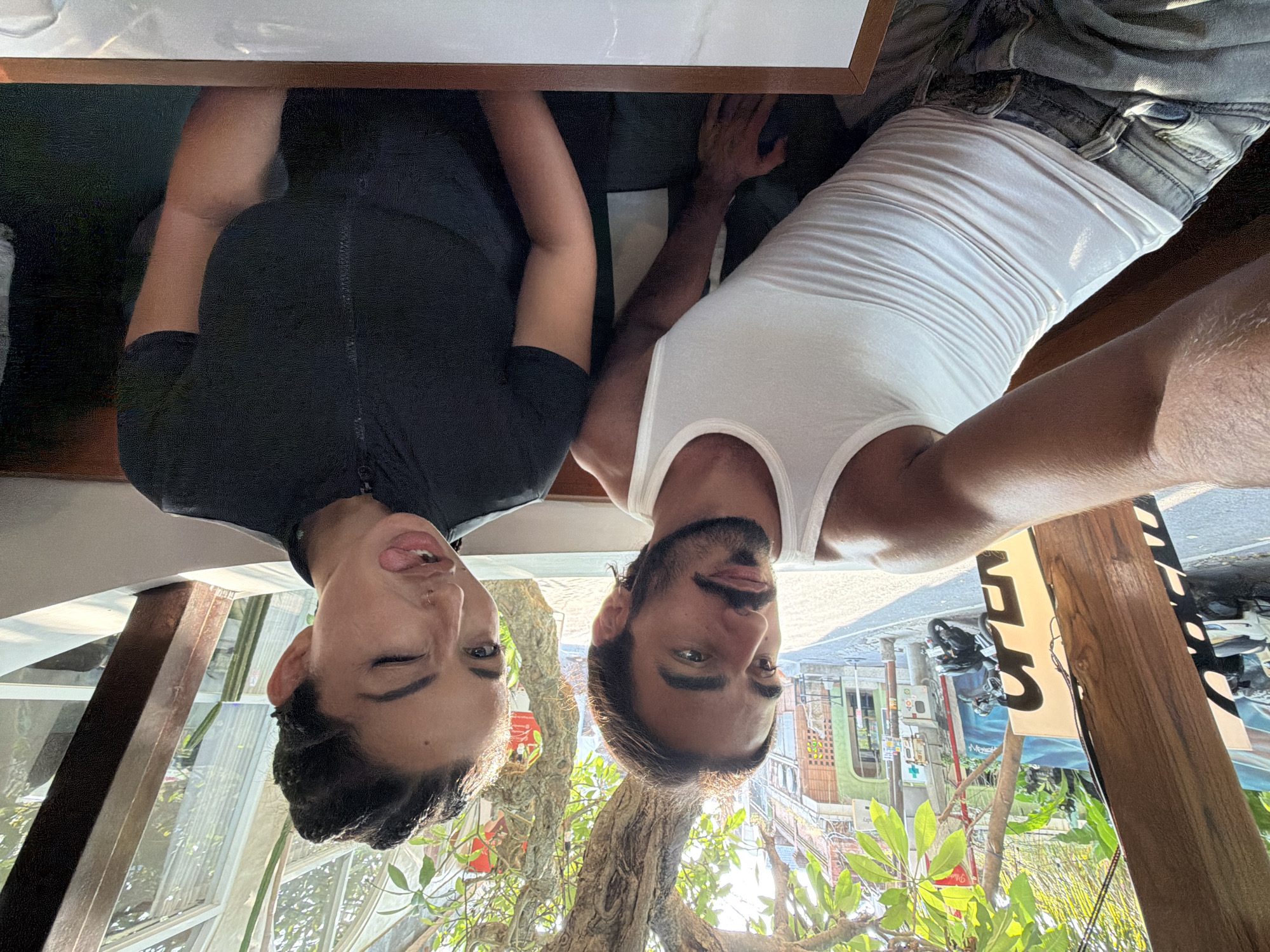 In Mengwi, Indonesia, a couple is captured in a selfie from the chest up, seated in a cafe. The man, on the left, wears a white tank top and light-wash jeans, his left arm resting on his lap. He has dark, shoulder-length hair, a mustache, and a beard, and he is looking to his left. The woman, on the right, has braided dark hair and is sticking her tongue out with one eye closed, looking directly at the camera. She is wearing a black short-sleeved zippered top. Behind them, through a wooden-framed window, a street scene unfolds with buildings, trees, and a parked motorcycle. The sunlight casts shadows, indicating it's daytime.
