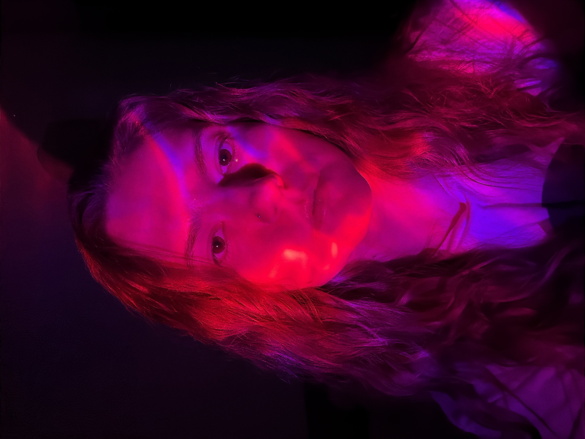 A close-up portrait shows a fair-skinned person with long, wavy hair under red and purple lighting. The person faces the camera with a neutral expression. The background is dark. No location-specific details are visible.