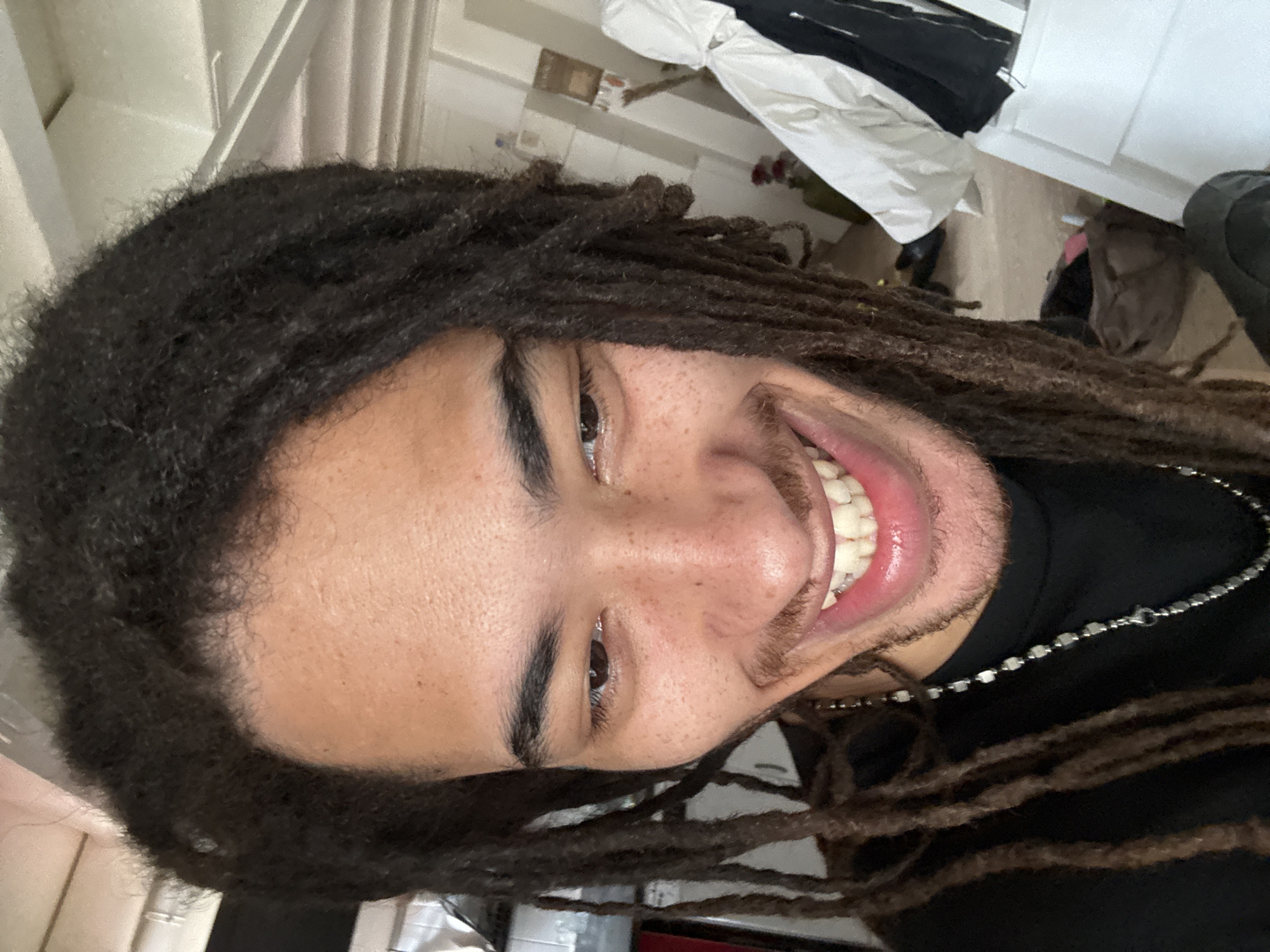 A person with dark dreadlocks, brown eyes, and a smile is facing the camera. The person is wearing a black shirt and a silver chain. The background appears to be an indoor space with various objects, including what seems like clothing, boxes, and furniture. There is no visual information in the image to confirm the location as Paris, France.